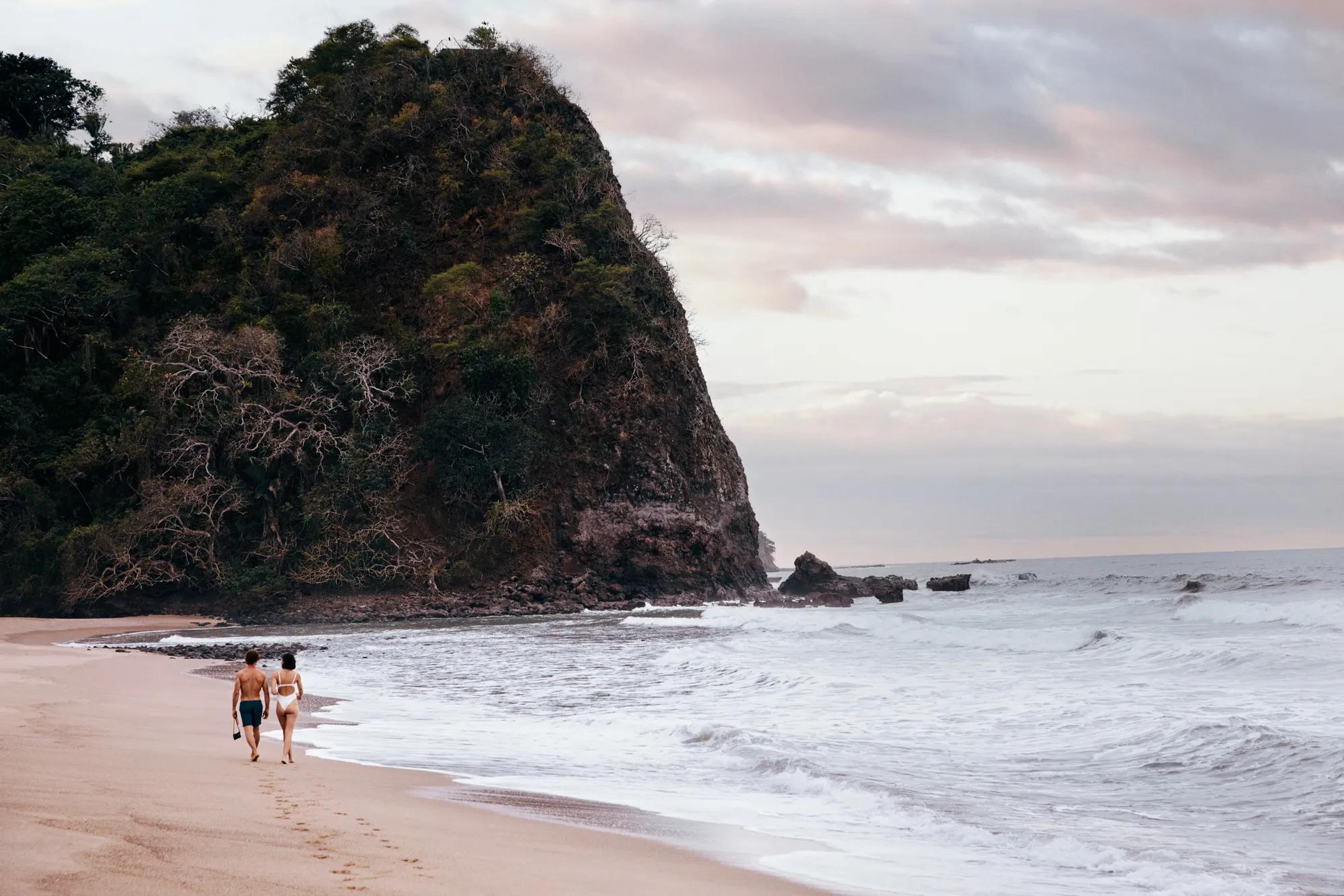 A couple walks barefoot along a secluded beach at sunset, with waves gently lapping the shore and a large, rocky cliff covered in lush greenery rising in the background.
