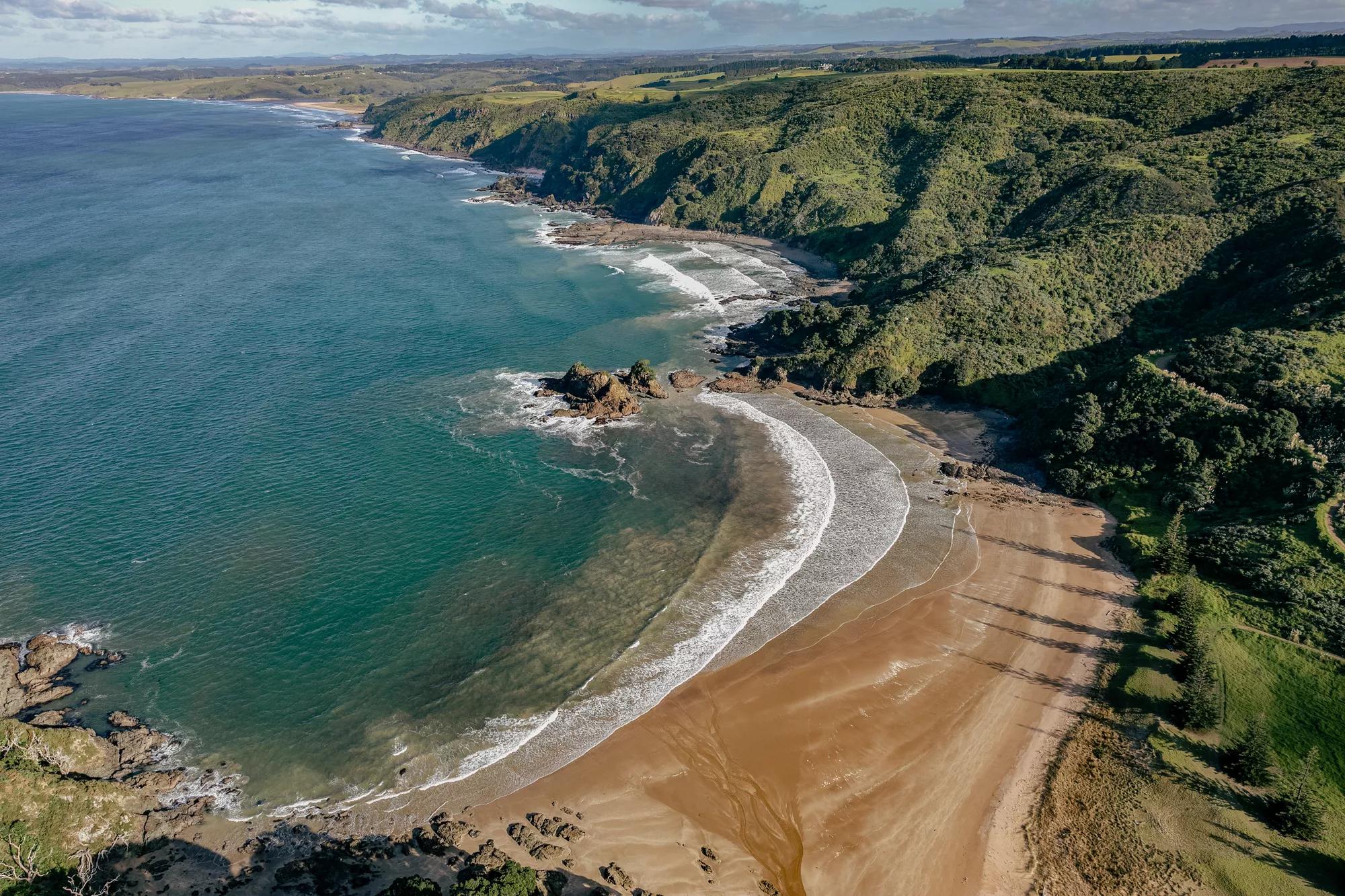 Aerial view of a white sand beach with clear blue waters and surrounded by lush green bush. 