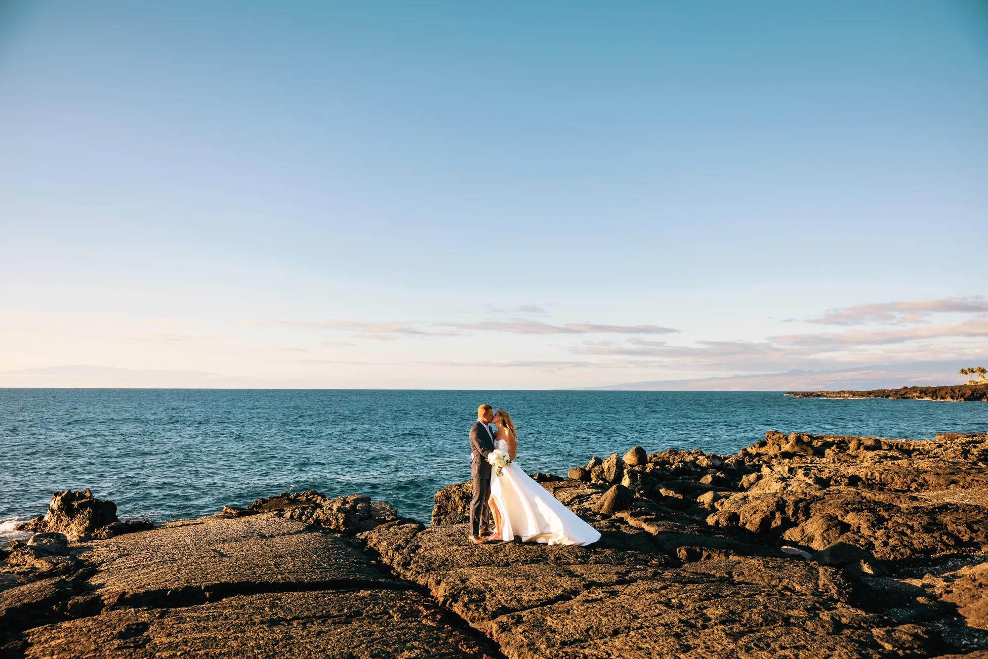 A bride and groom embracing on lava rock against a backdrop of the ocean