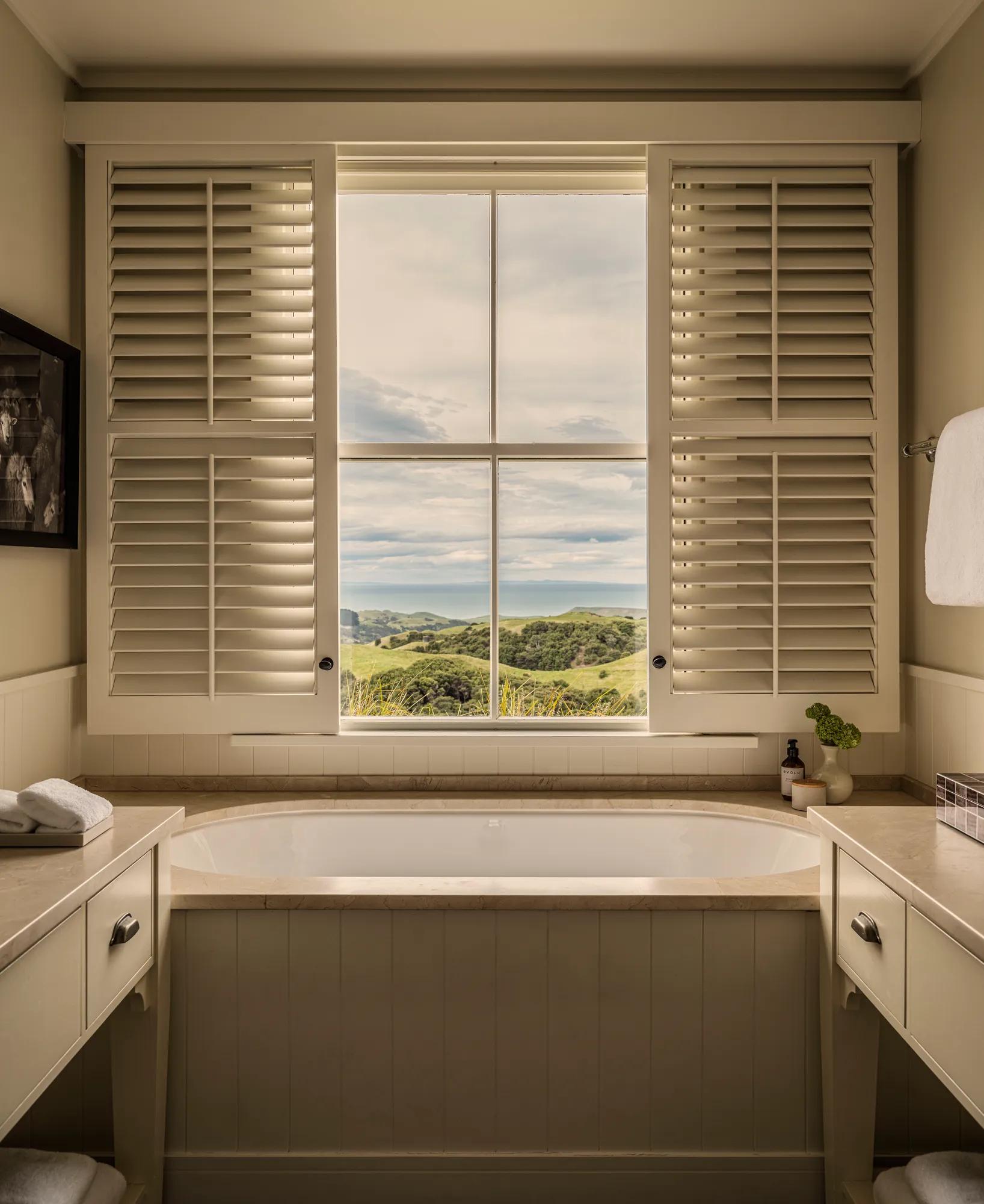 Oversized bathtub in a hotel room with the window open behind open shutter revealing views of rolling farmland.