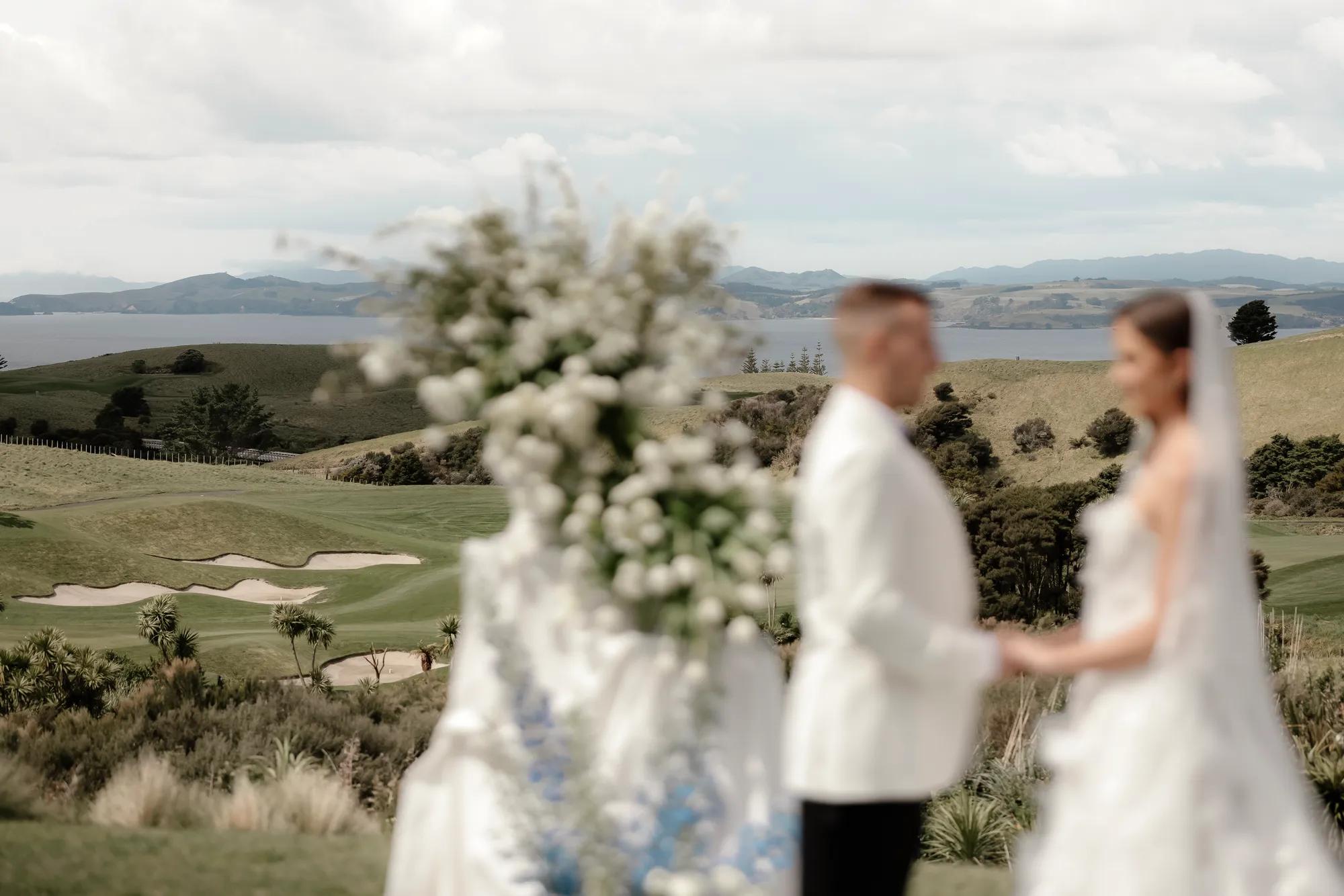 Bride and groom at the alter, blurred in the foreground, with a vast landscape and golf course view in focus in the background. 