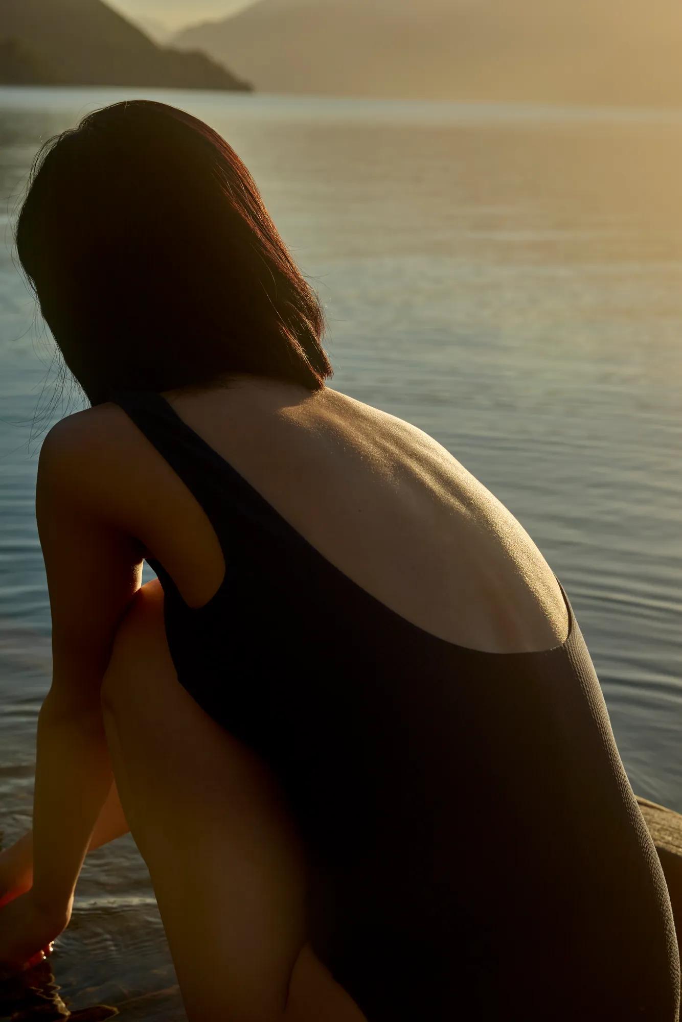 Back view of a woman crouched beside Lake Wakatipu in a swimsuit looking out to a serene view of the lake and mountains as the sun sets.