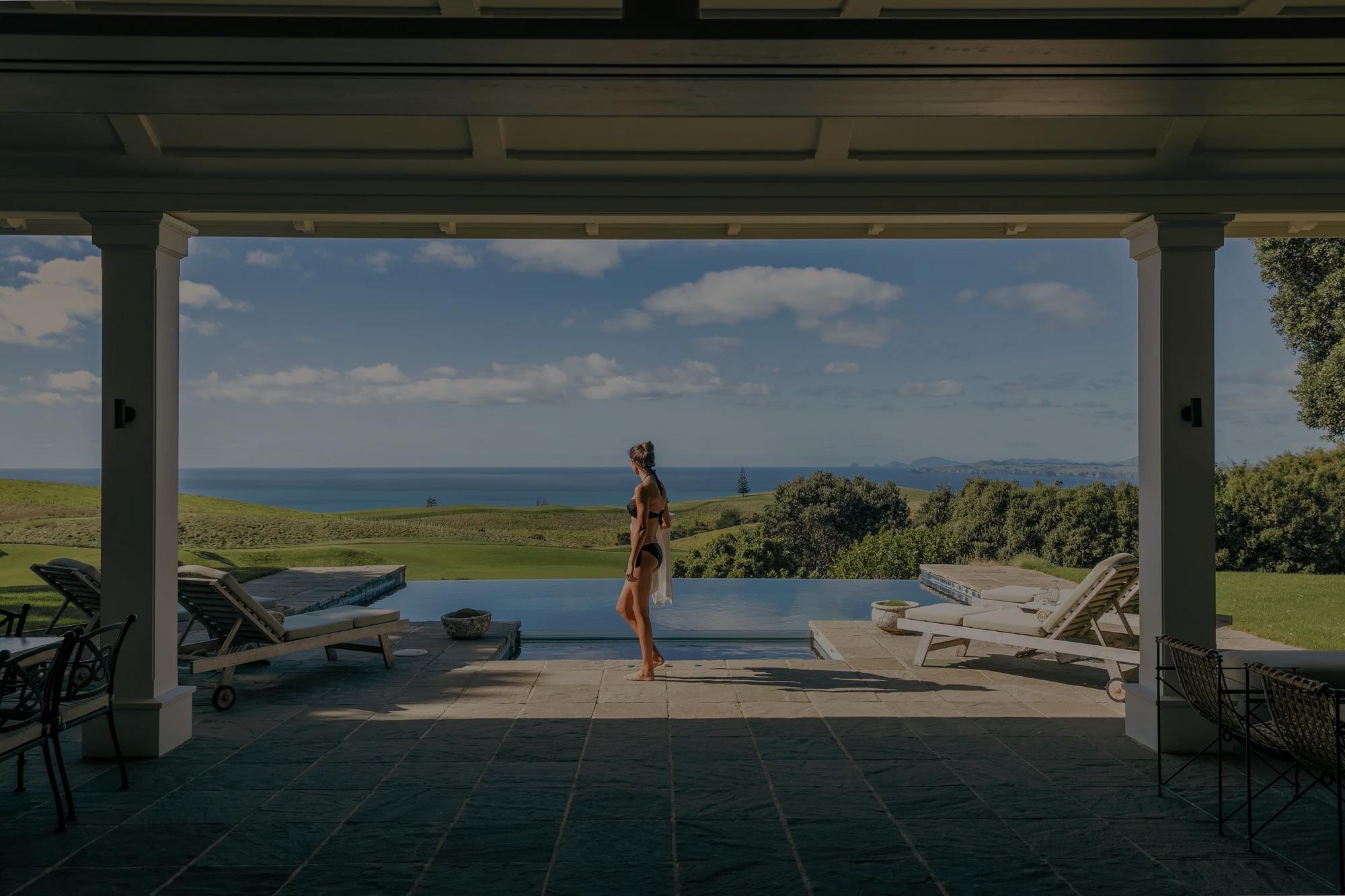 Woman in a bikini walking in front of the outdoor pool in the Kauri Cliffs Two Bedroom Villa, she is carrying a towel over her shoulder and looking out to the views of the golf course and Pacific Ocean.