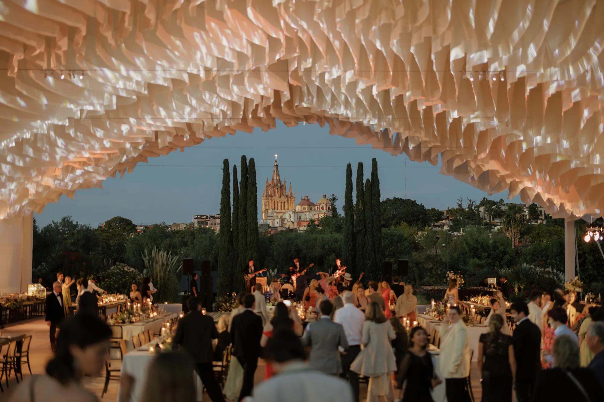 Outdoor wedding reception with flowing fabric draped from the ceiling structure, elegant setup beneath, and the Parroquia de San Miguel Arcángel in the background.