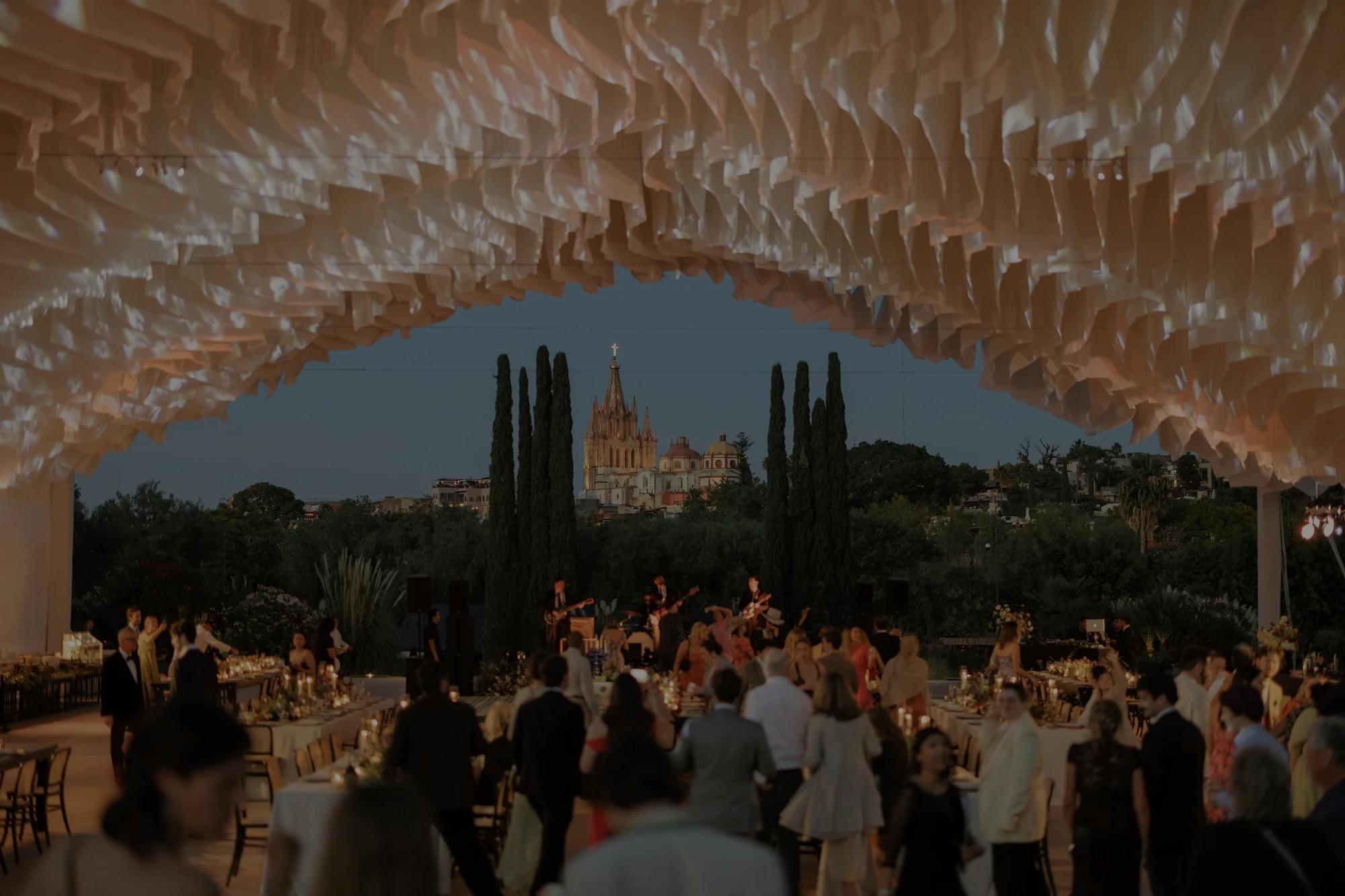 Event setup at Jardín Rosewood with guests gathered under a draped canopy, set on a garden lawn with the Parroquia de San Miguel Arcángel in the background.