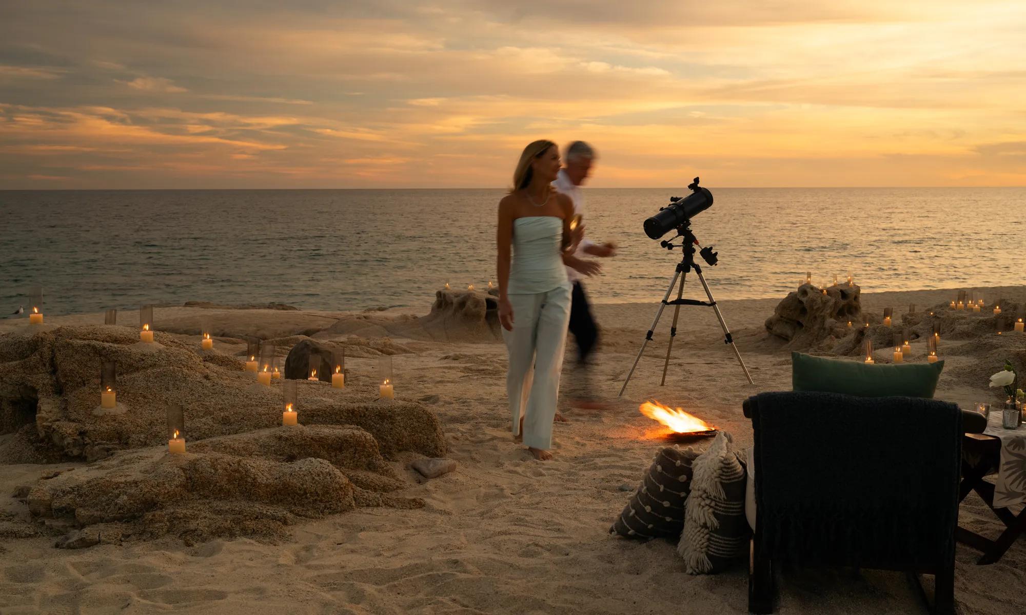 Couple walking along a candlelit beach at sunset in Los Cabos, with a telescope and intimate seaside setup creating a romantic starlit experience by the ocean.