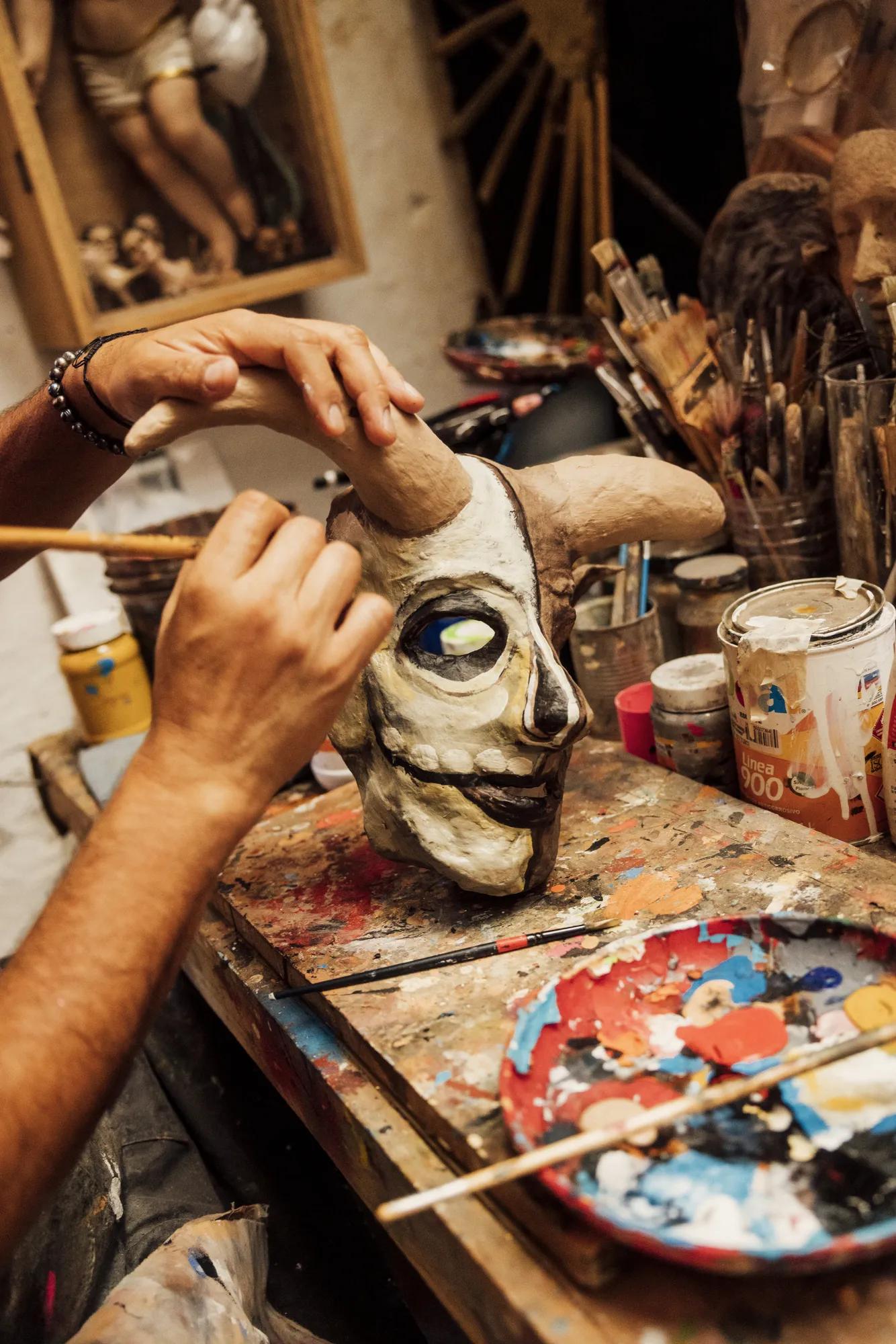 Artist’s hands painting a colorful mask on a table, surrounded by various paint supplies and brushes in a creative art studio.