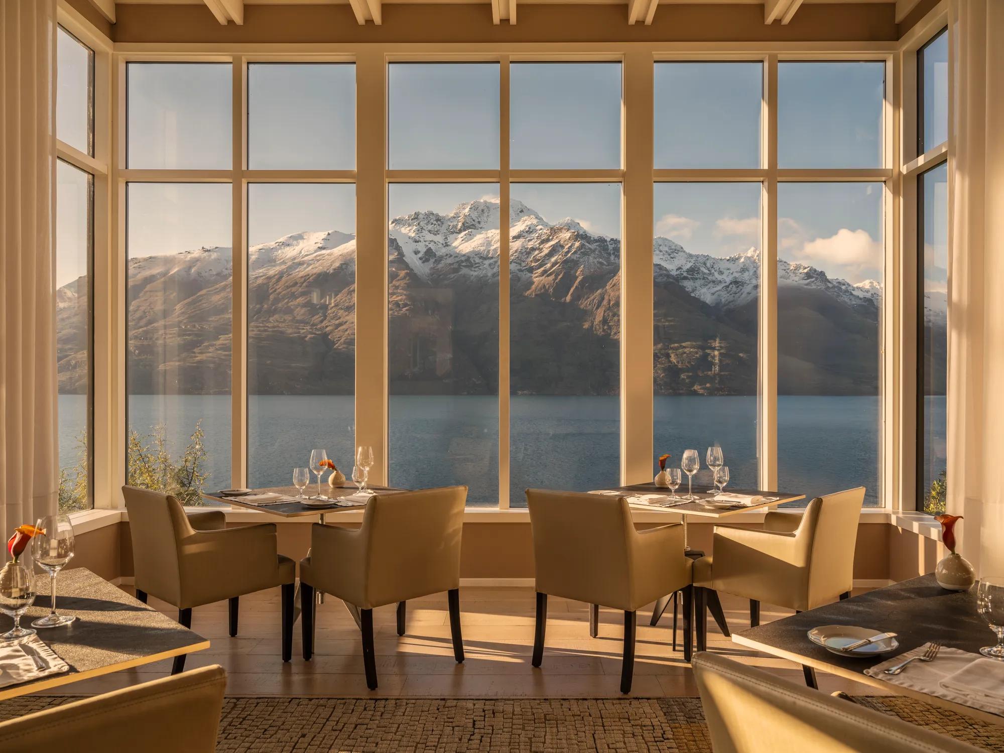Four set tables in the dining room at Rosewood Matakauri, two tables are the focus and are positioned next to the big picture windows with views of Lake Wakatipu and snow-capped mountains.