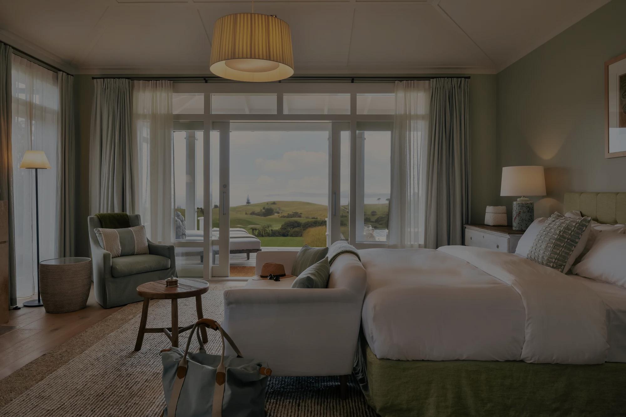 Primary bedroom in the Kauri Cliffs Four Bedroom Villa with luxurious bed, living room with stylish furnishing and opening doors to the deck and views of the Pacific Ocean beyond.