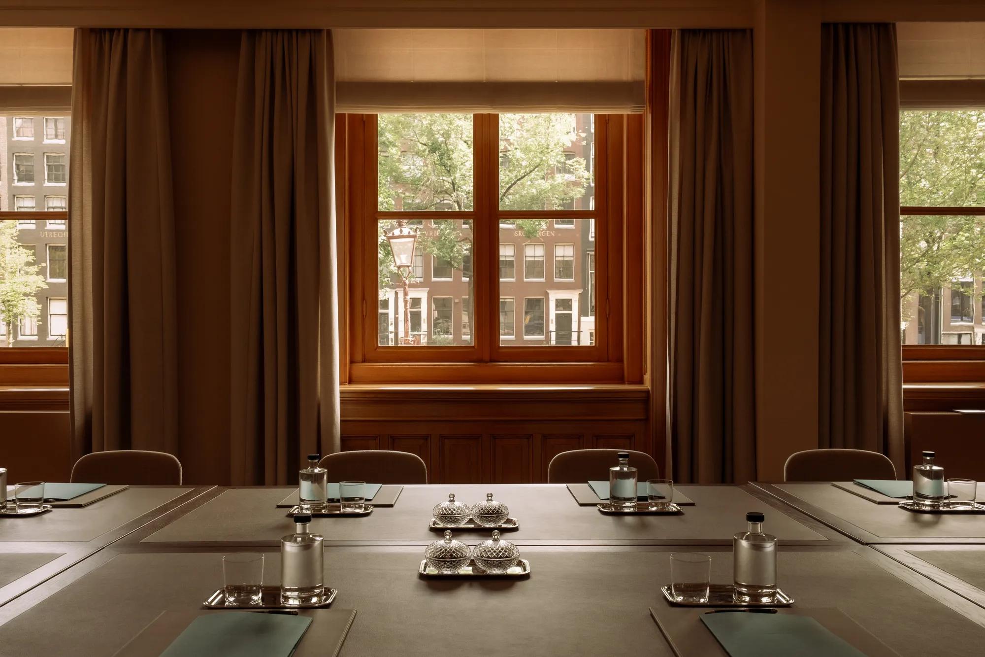 Tables set up for a boardroom meeting, looking out on brick canal houses, trees and a lamppost. Wooden pannelling and drapery frame the windows. 