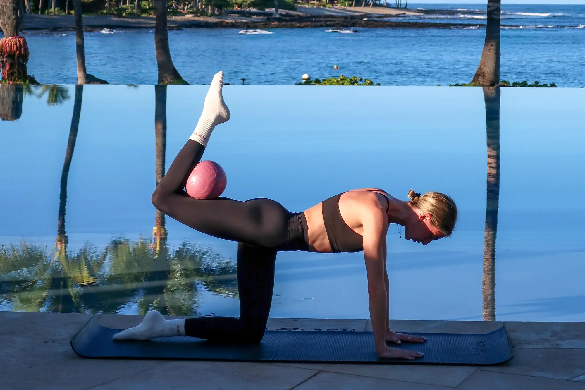Person performing a Pilates exercise on a mat by an infinity pool, balancing a pink ball behind the knee with the ocean in the background.