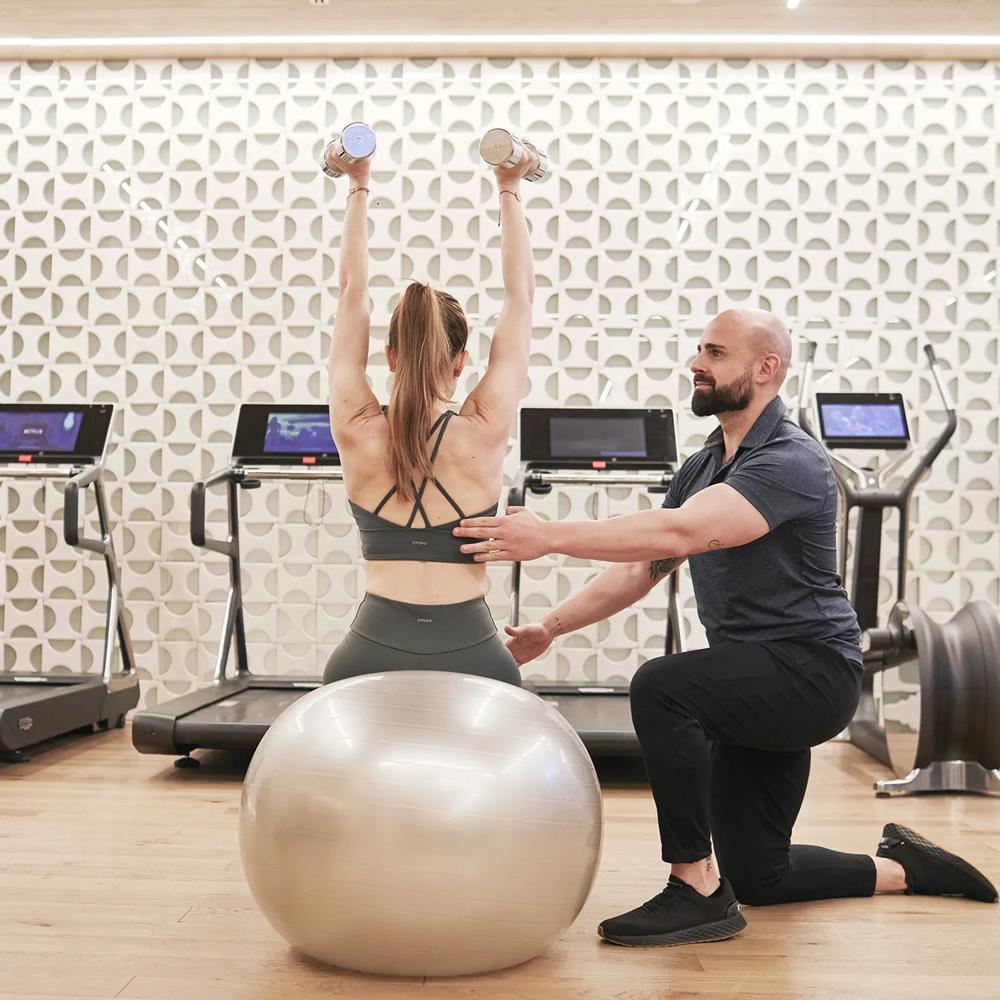 Girl in fitness attire seated on a gym ball, assisted by a personal trainer who is guiding and correcting her posture in a modern fitness area