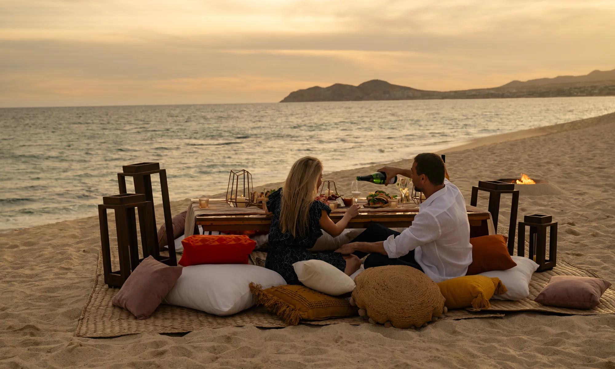 Couple enjoying a sunset beach picnic in Los Cabos, seated on plush cushions with lanterns, ocean views, and a curated spread of food and wine by the Sea of Cortez.