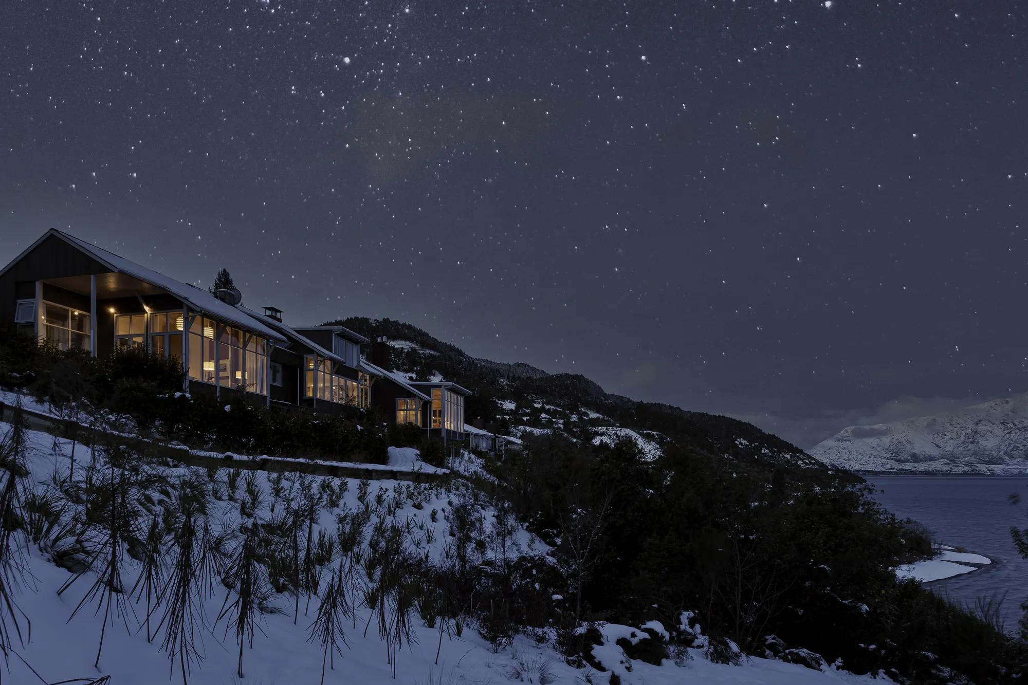 Exterior low angle view of the Matakauri lodge on a snowy evening with the starry night sky visible in the background. 