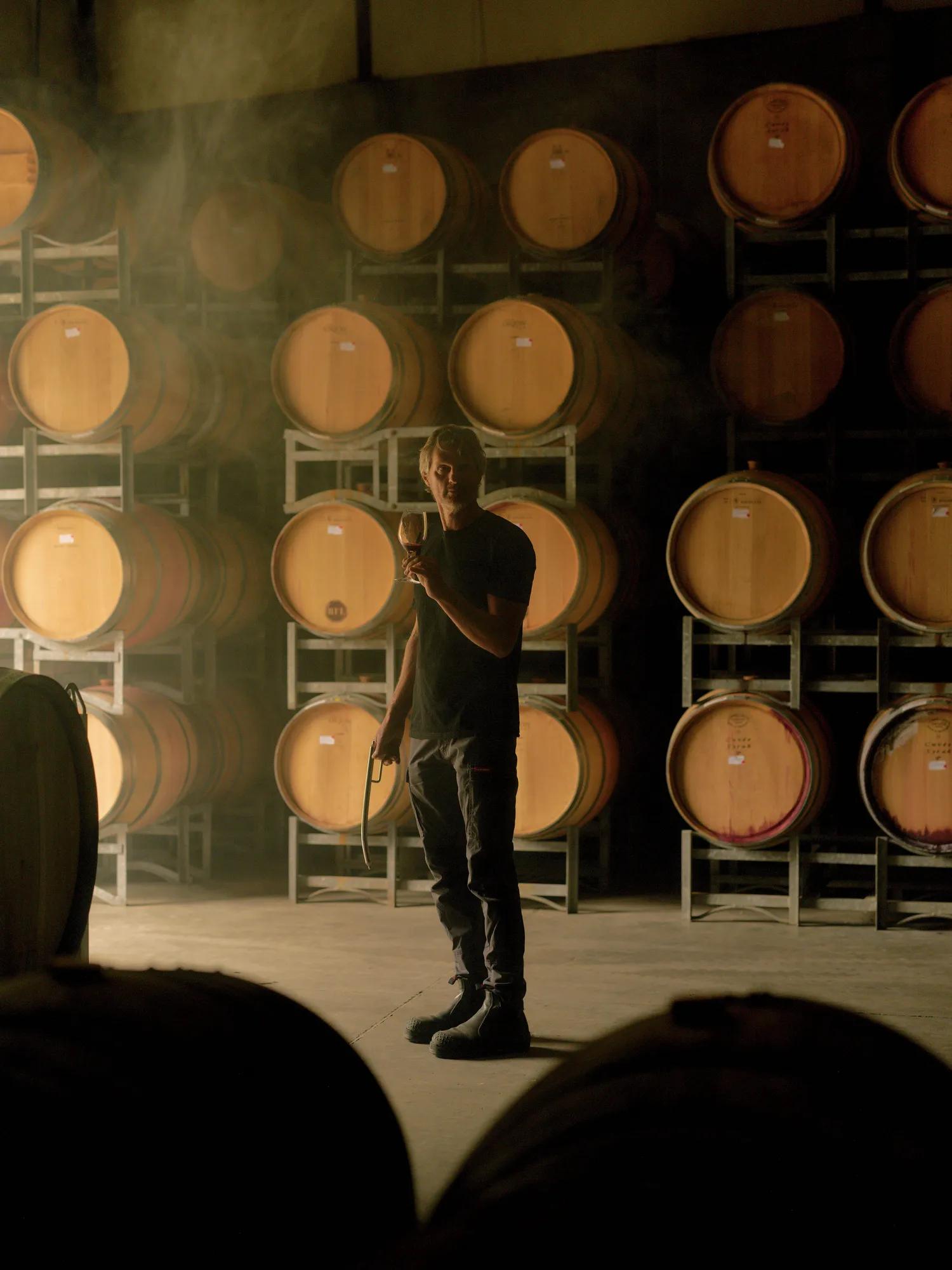 Man holding wine glass in a cellar with wooden barrels stacked on racks.