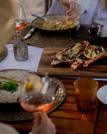 A beachside dining table with assorted dishes and wine and a wooden deck overlooking a sandy shore.