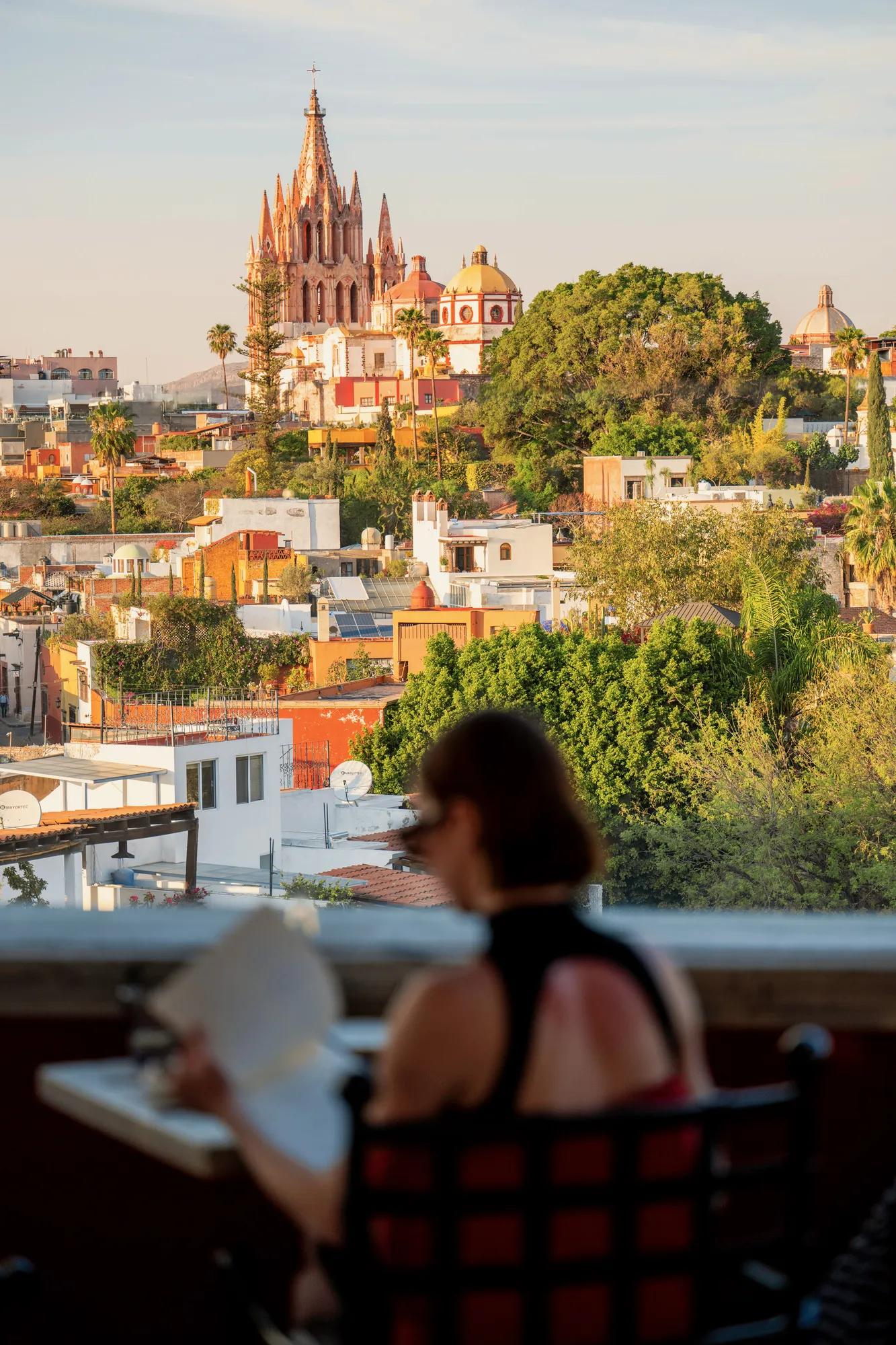 Woman seated at Luna Rooftop reading the menu, with views of the Parroquia, colorful houses, and treetops of San Miguel de Allende in the background.