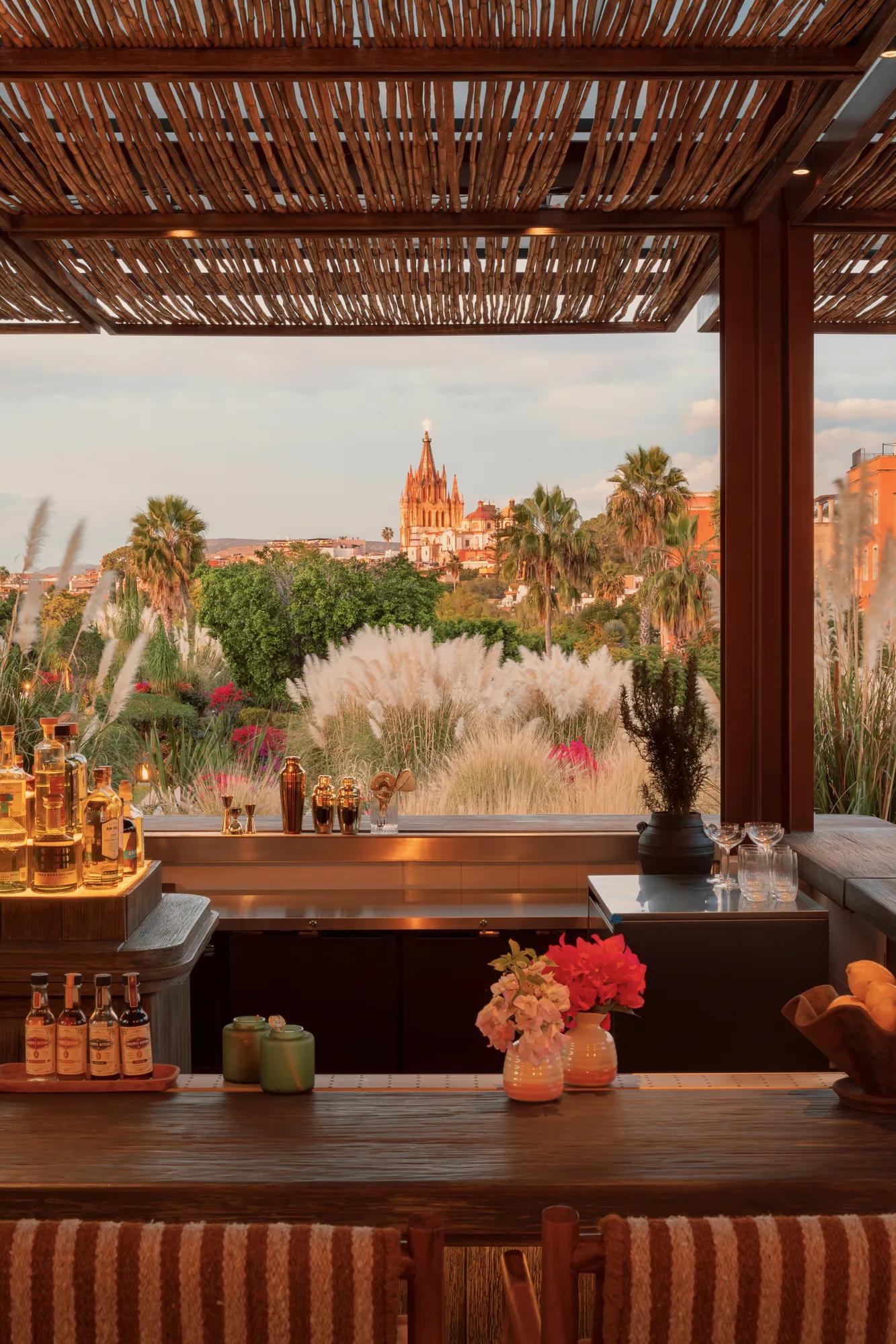 An open-air bar with bottles displayed on a backlit shelf, overlooking the hotel gardens and the Parroquia in the distance. 