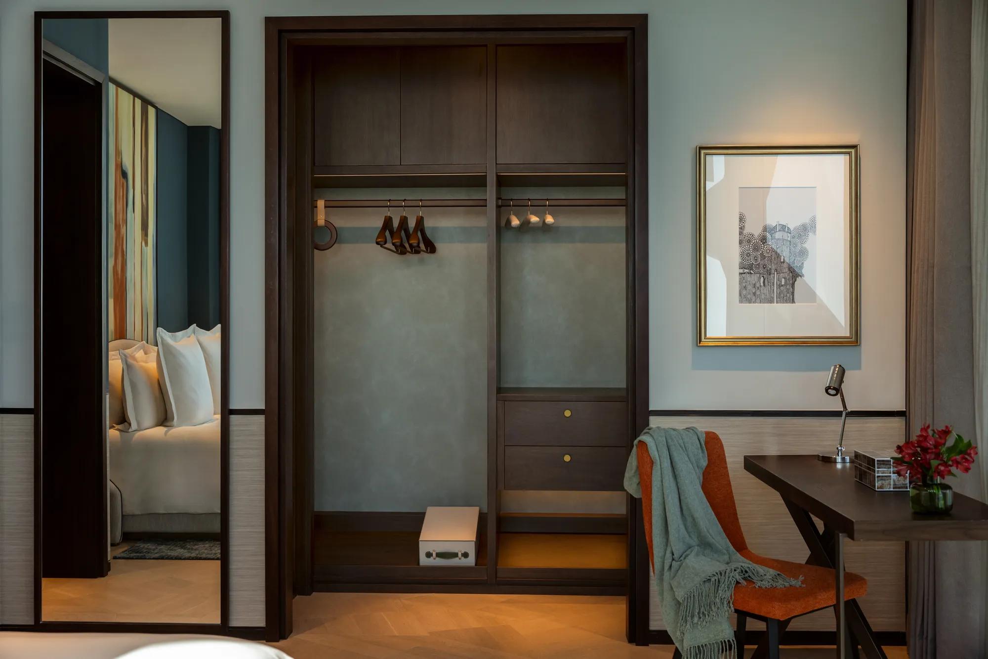 Elegant hotel room interior featuring a built-in wardrobe with hangers, a desk with an orange chair, and a partial view of the bedroom through a mirror.