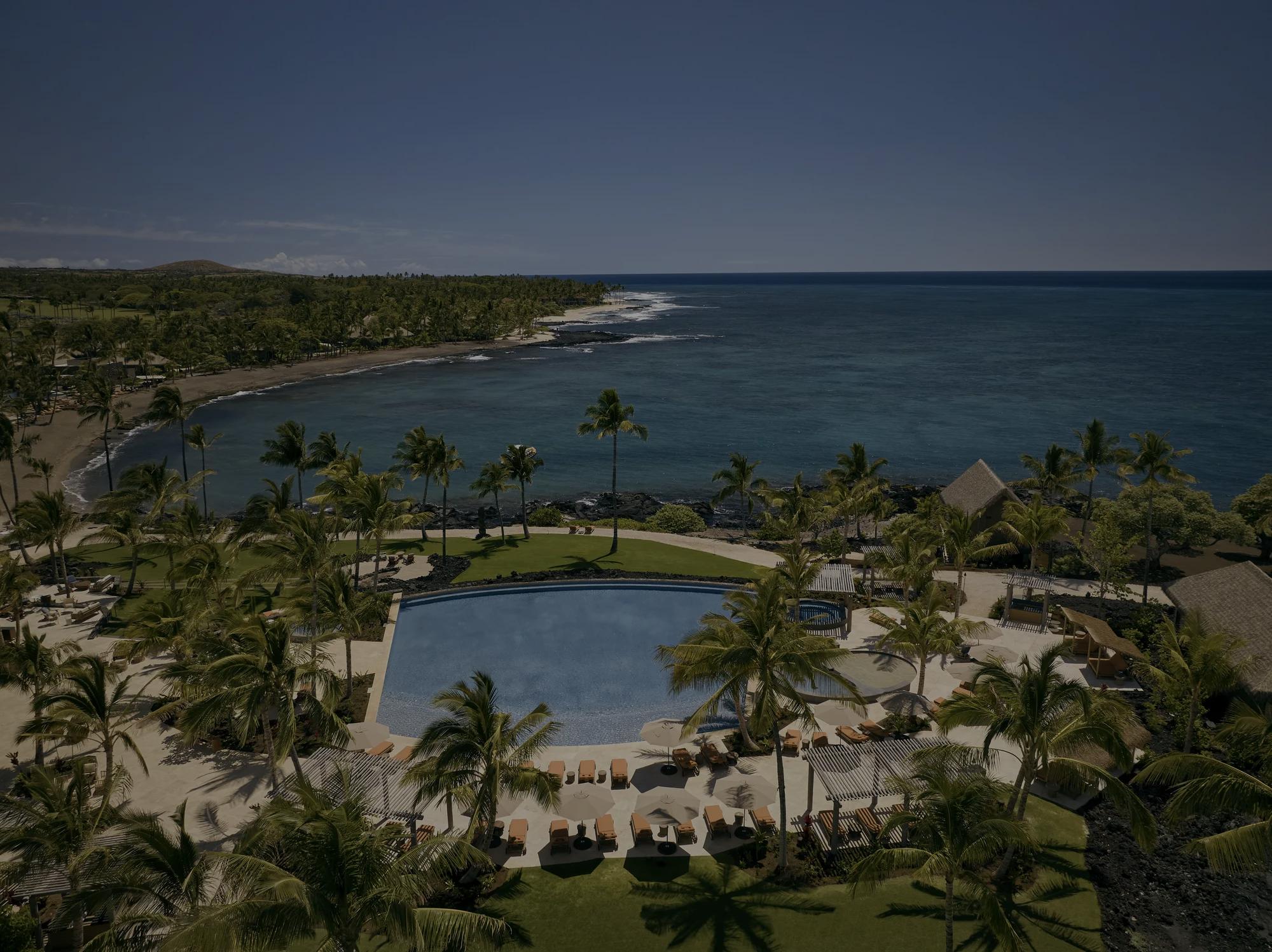 Aerial drone shot of an ocean front pool overlooking a sparkling blue bay with sprawling palms surrounding the pool with the Kona coastline in the distance
