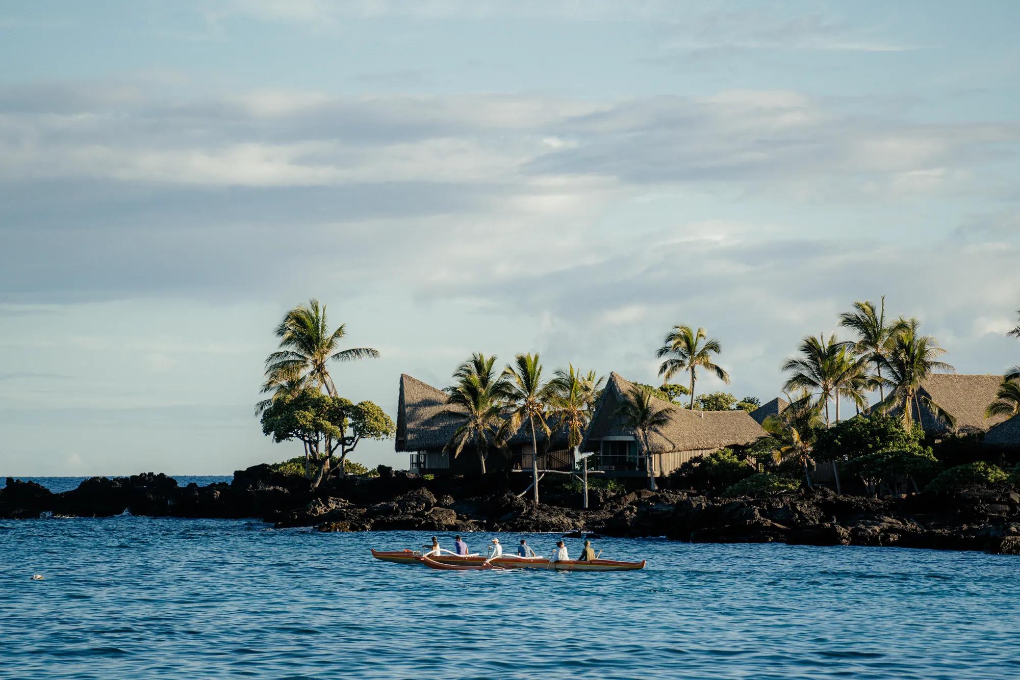 People paddling on a canoe in the ocean with thatched roof bungalows and plan trees on the volcanic rock shoreline in the background.
