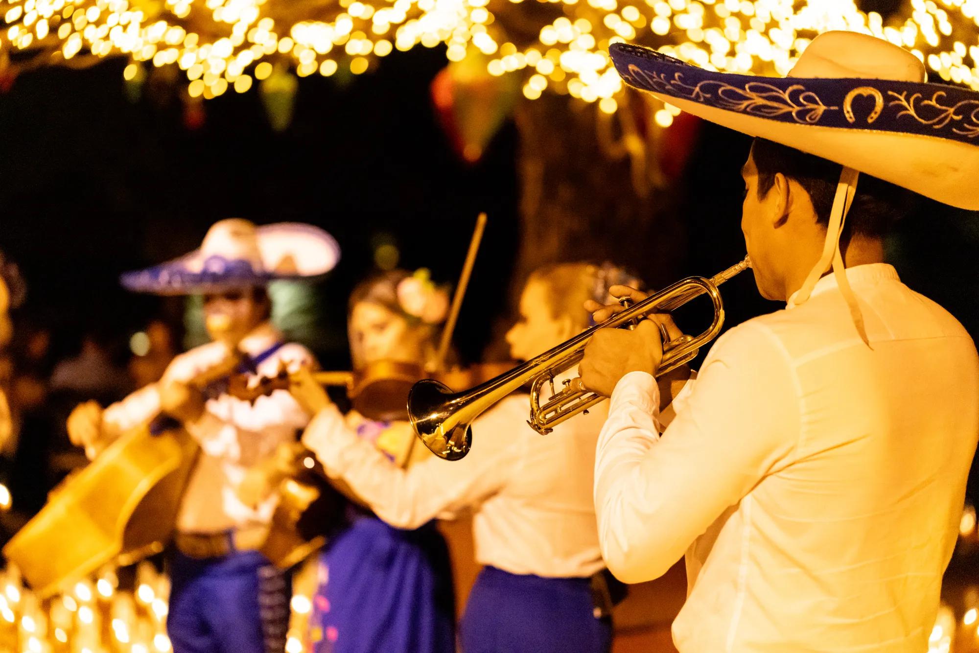A woman in traditional folkloric dress dances gracefully, holding a decorative fan and lifting her lace skirt, under glowing golden lights. A man in a white outfit and hat stands smiling in the background, creating a festive Día de Muertos atmosphere.