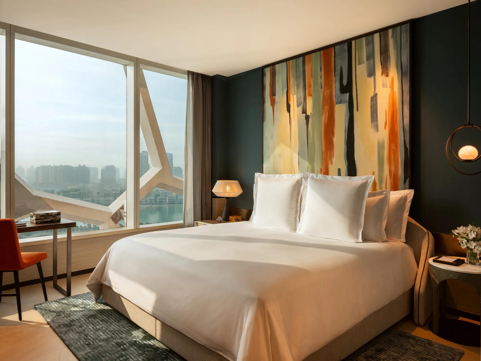 Contemporary hotel bedroom with large windows overlooking a city skyline, featuring crisp white bedding and an abstract art headboard in orange, blue, and beige tones.