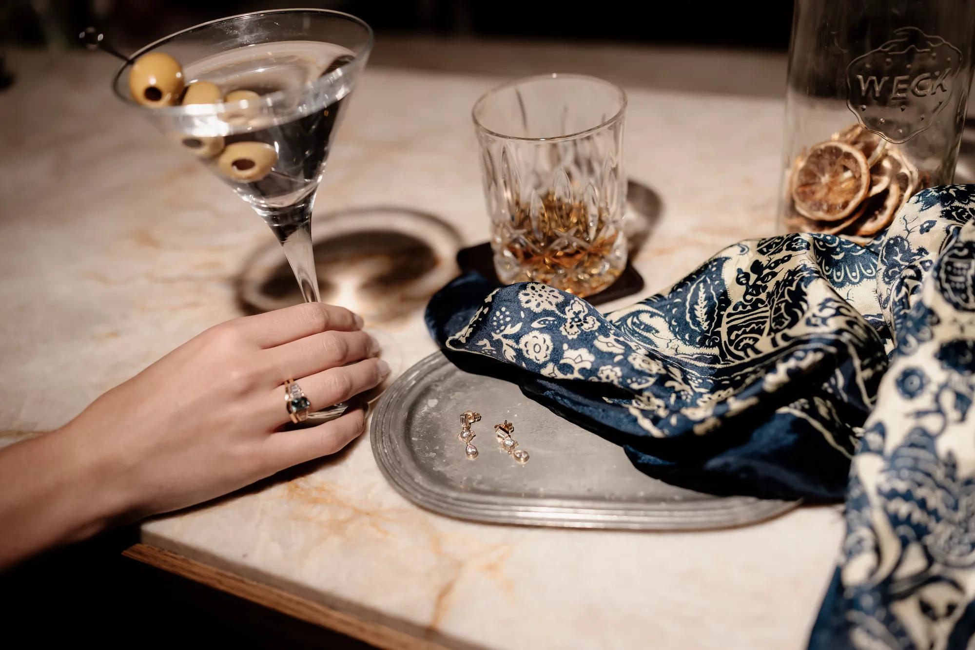 Martini and whisky cocktails on a bar counter, elegant female hand holding the base of a martini glass, silk scarf and metal tray with earrings displayed on bar surface.