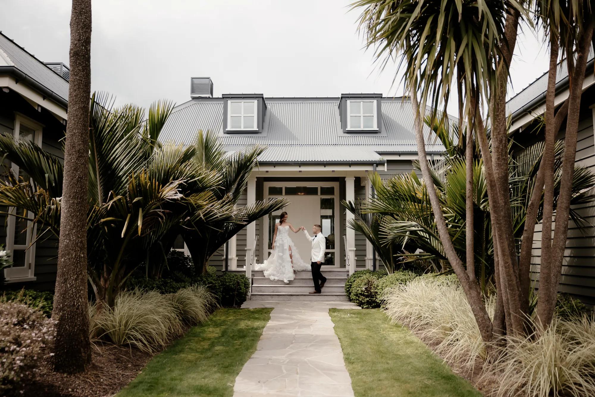 Groom leading bride down steps of luxury villa towards a stone path, surrounded by manicured gardens.