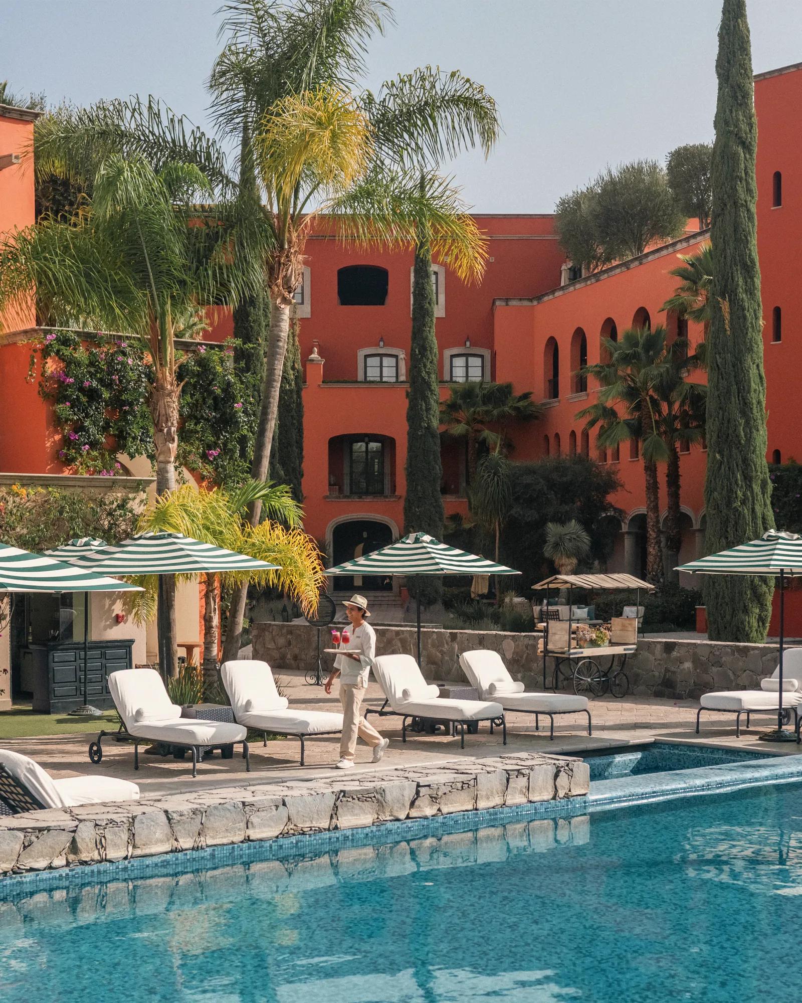 A waiter carries two glasses while walking by the poolside, surrounded by loungers, umbrellas, palm trees, and pines, with the hotel façade in the background.