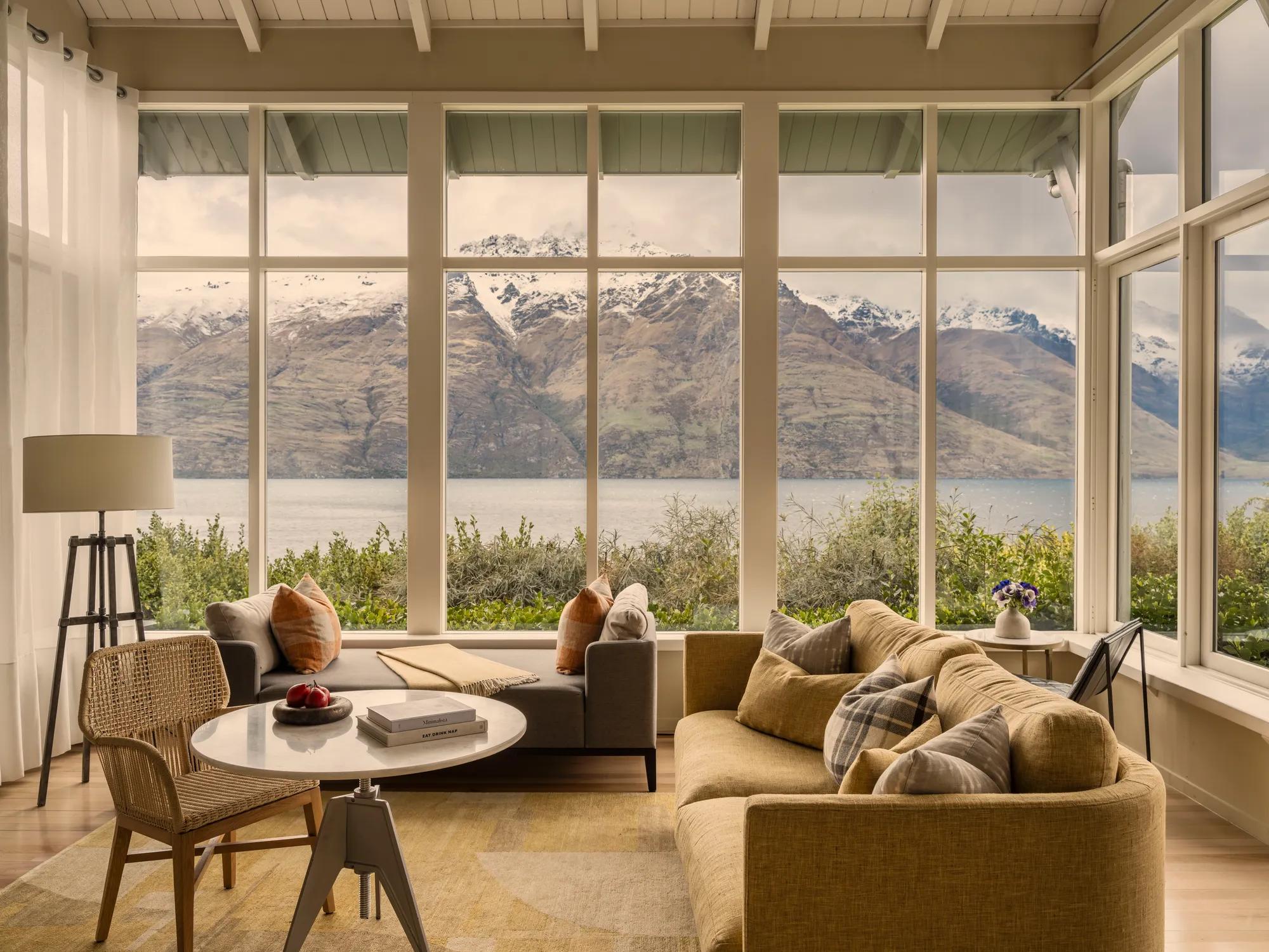 Hotel suite living area with neutral furnishings including a couch, day bed, table and chair, floor to ceiling windows that look out a lake and snowy mountains.