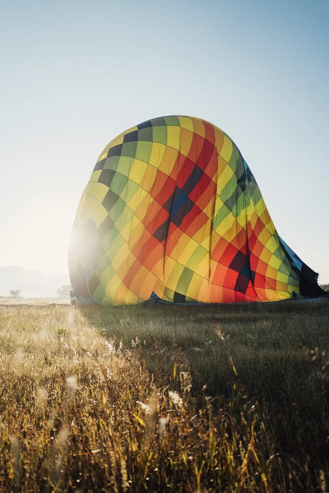A colorful hot air balloon partially inflated and resting on the grass, with four people holding it steady under a clear sky in an open field.