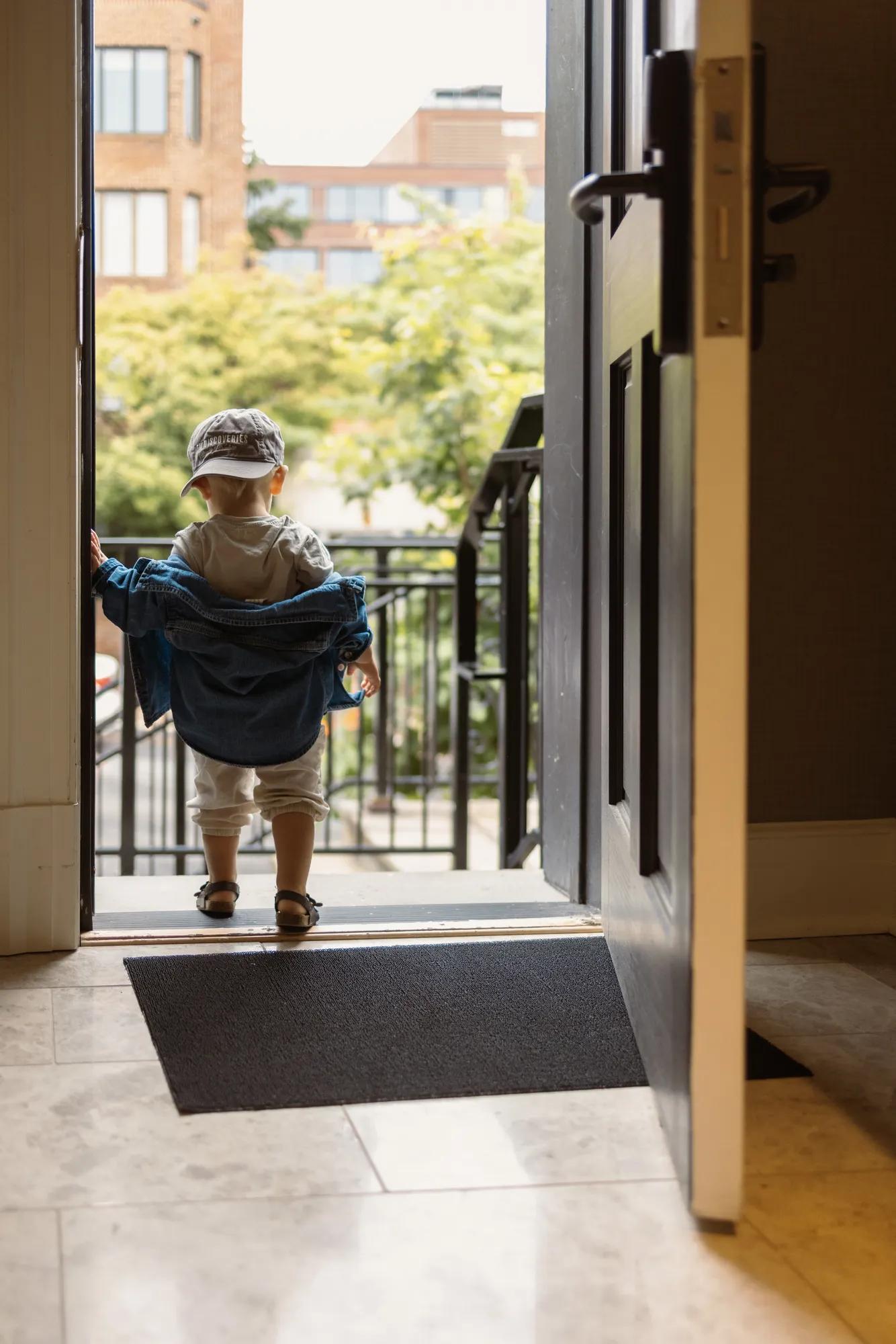 Child in doorway of townhouse heading out to the sidewalk with a backwards hat on.