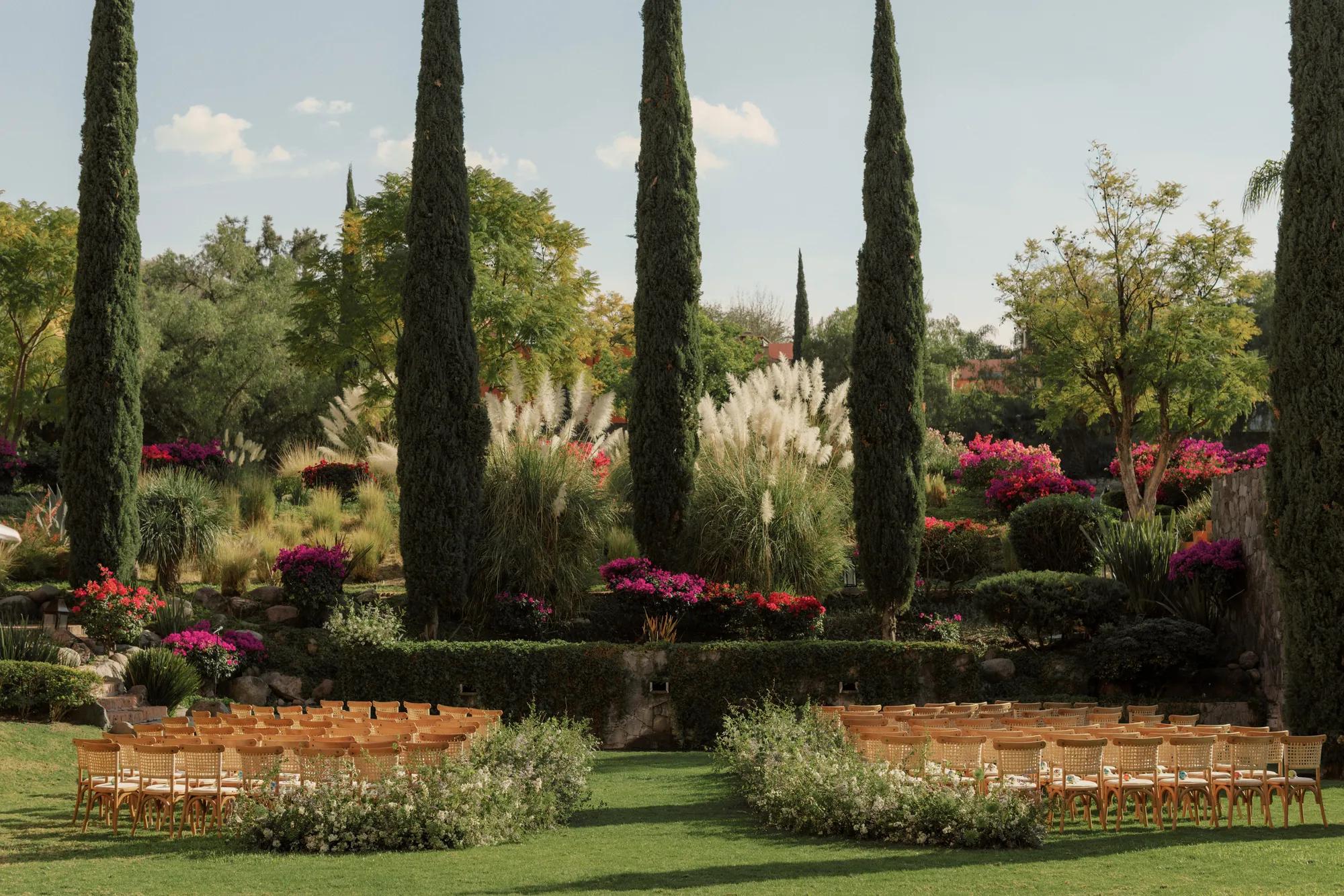 Garden wedding ceremony setup with a central aisle lined with floral arrangements and rows of chairs on each side, surrounded by lush greenery.