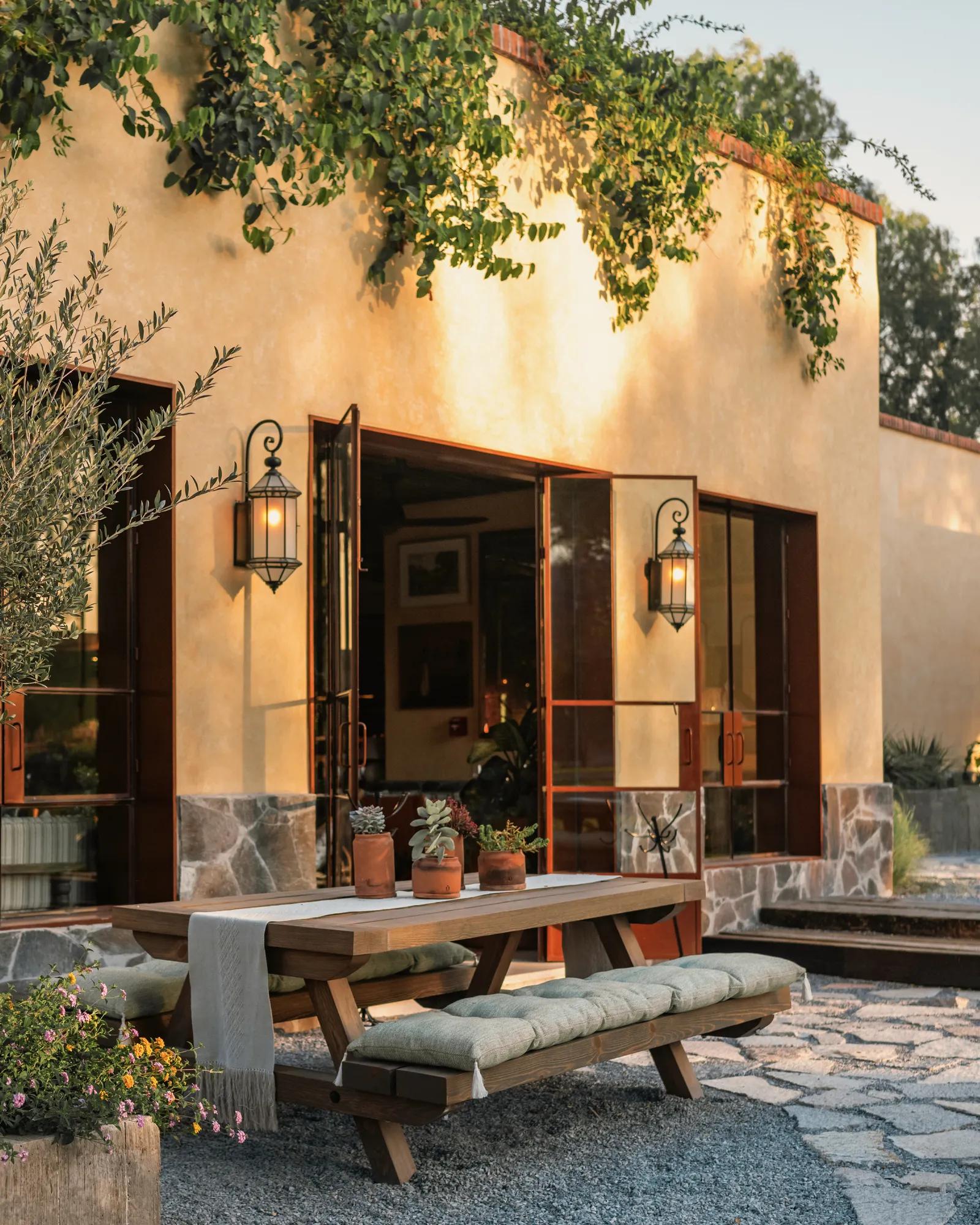 A young woman wearing a hat, white shirt, and light jeans rides a brown horse along a trail with two other riders ahead and behind, surrounded by dry trees.
