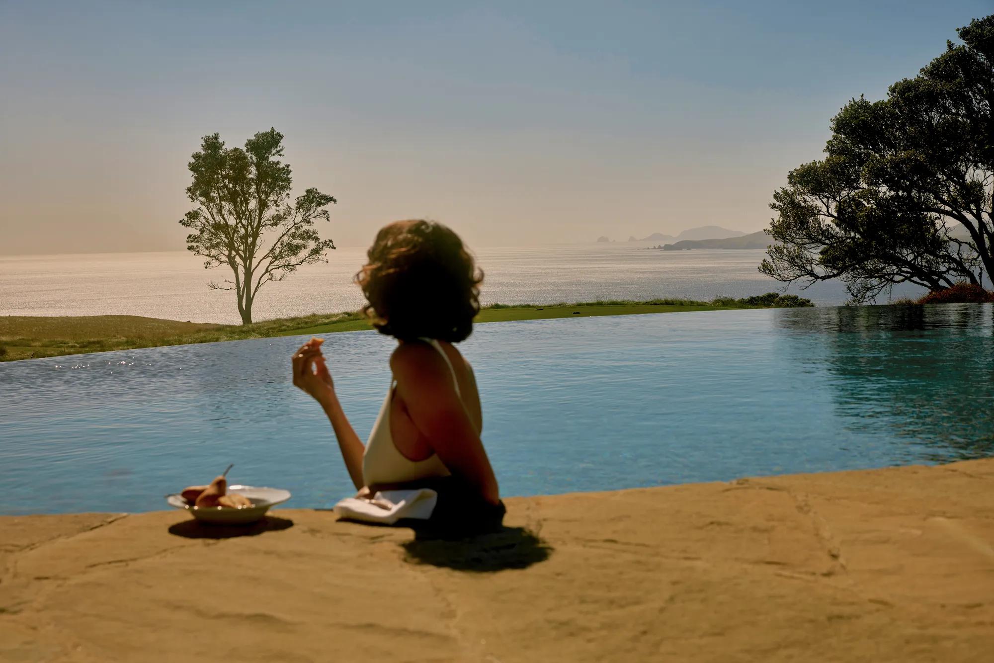 Woman leans on the edge of an infinity pool eating a slice of Pear as she looks out at views of the golf course and the Pacific Ocean.