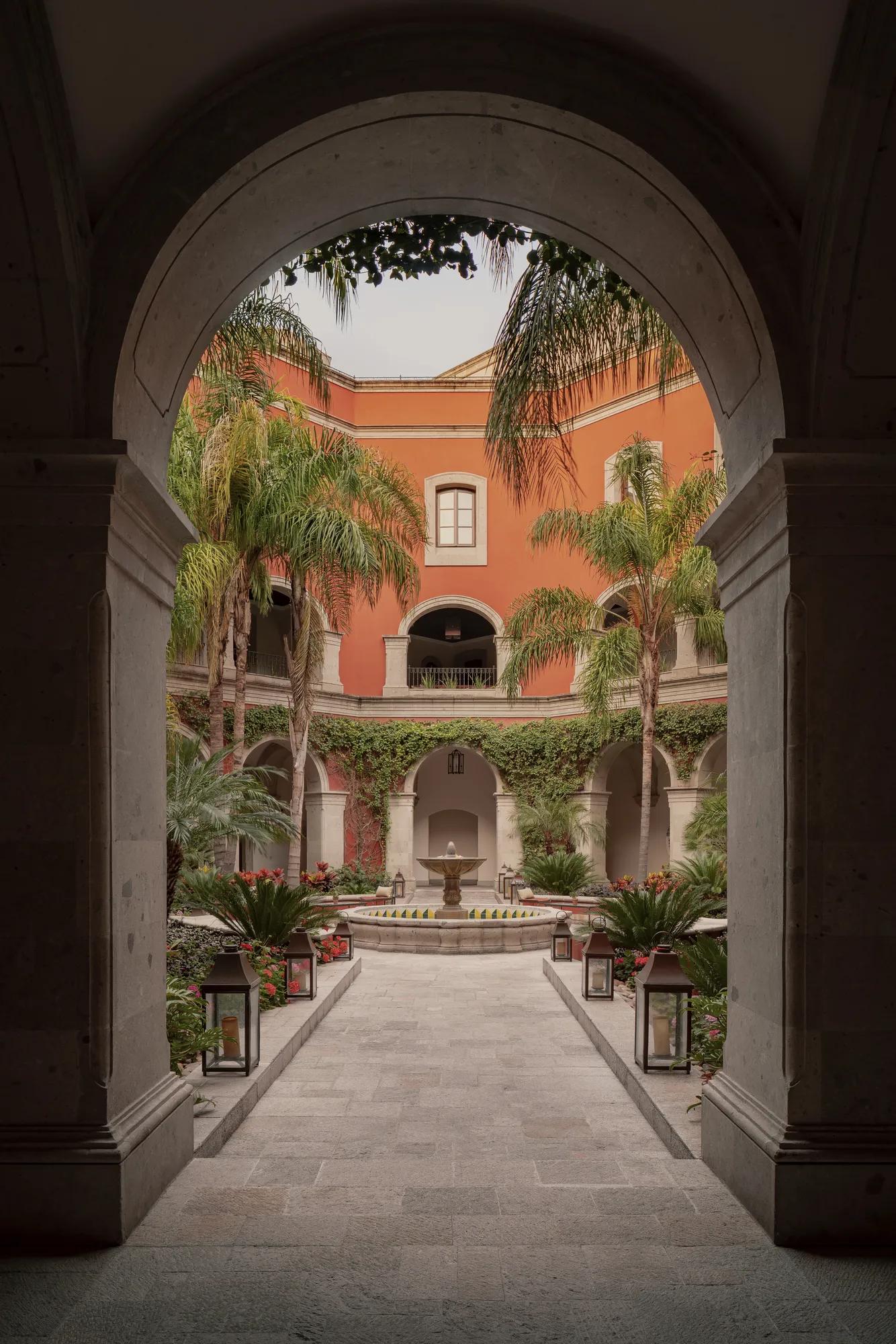 A central stone fountain in a courtyard, surrounded by a path with floor lights and bordered by planters with lush greenery and palm trees. 