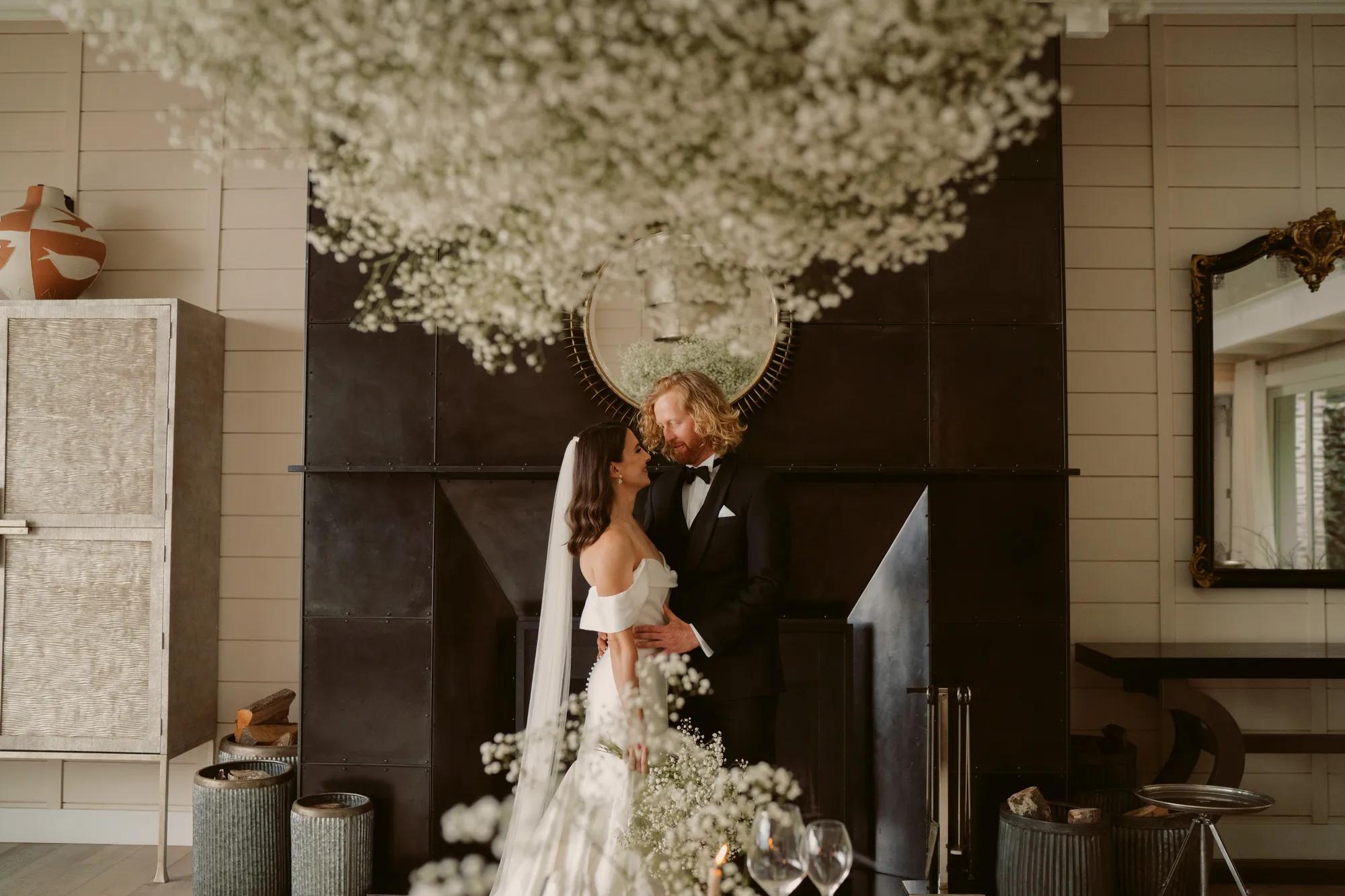 Bride and Groom stand in front of a fireplace in a luxury villa at Rosewood Matakauri, simple white floral installation above them. 