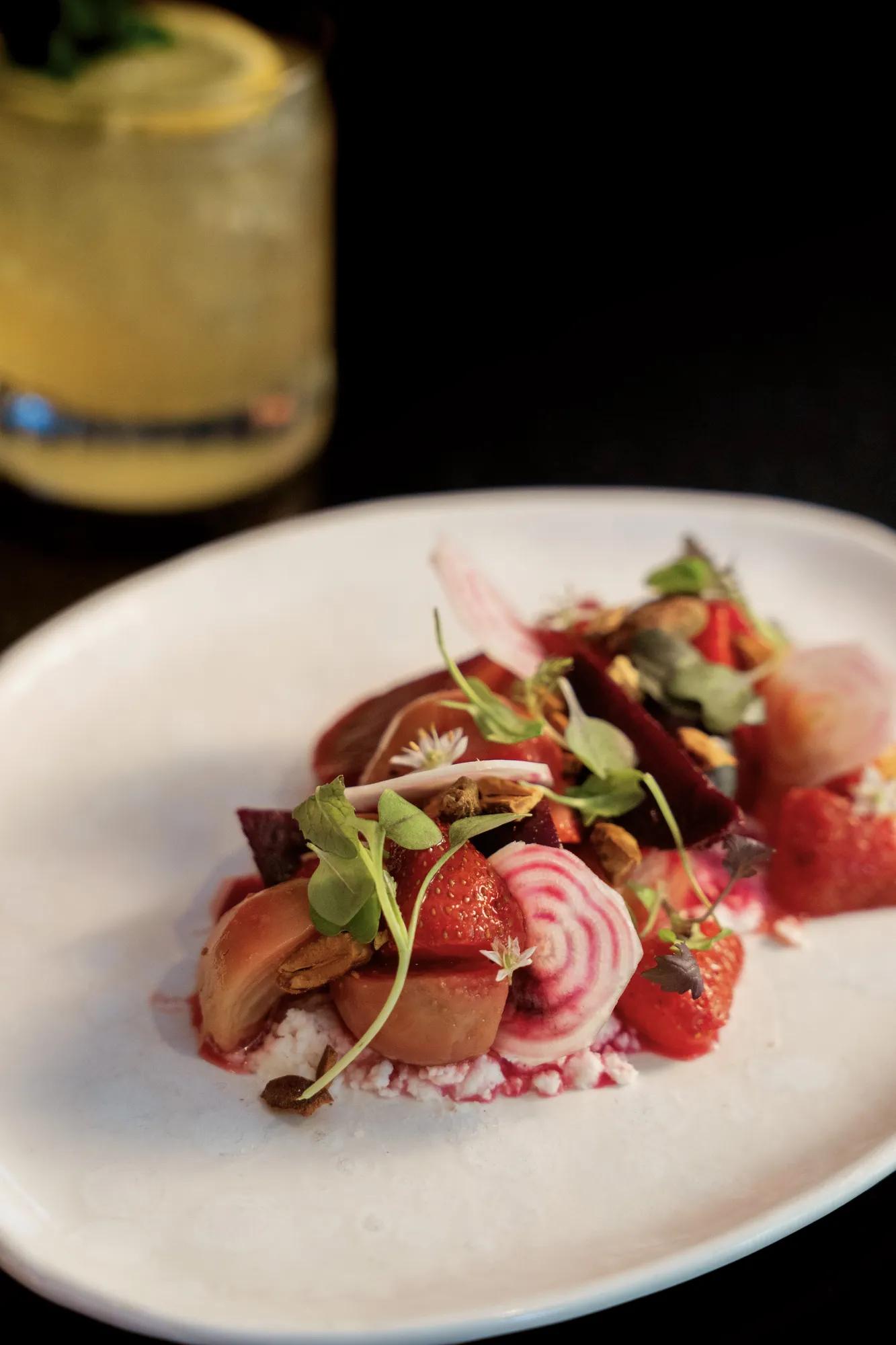 Strawberry and beet salad with goat cheese on the bottom on a white plate with a cocktail in the background