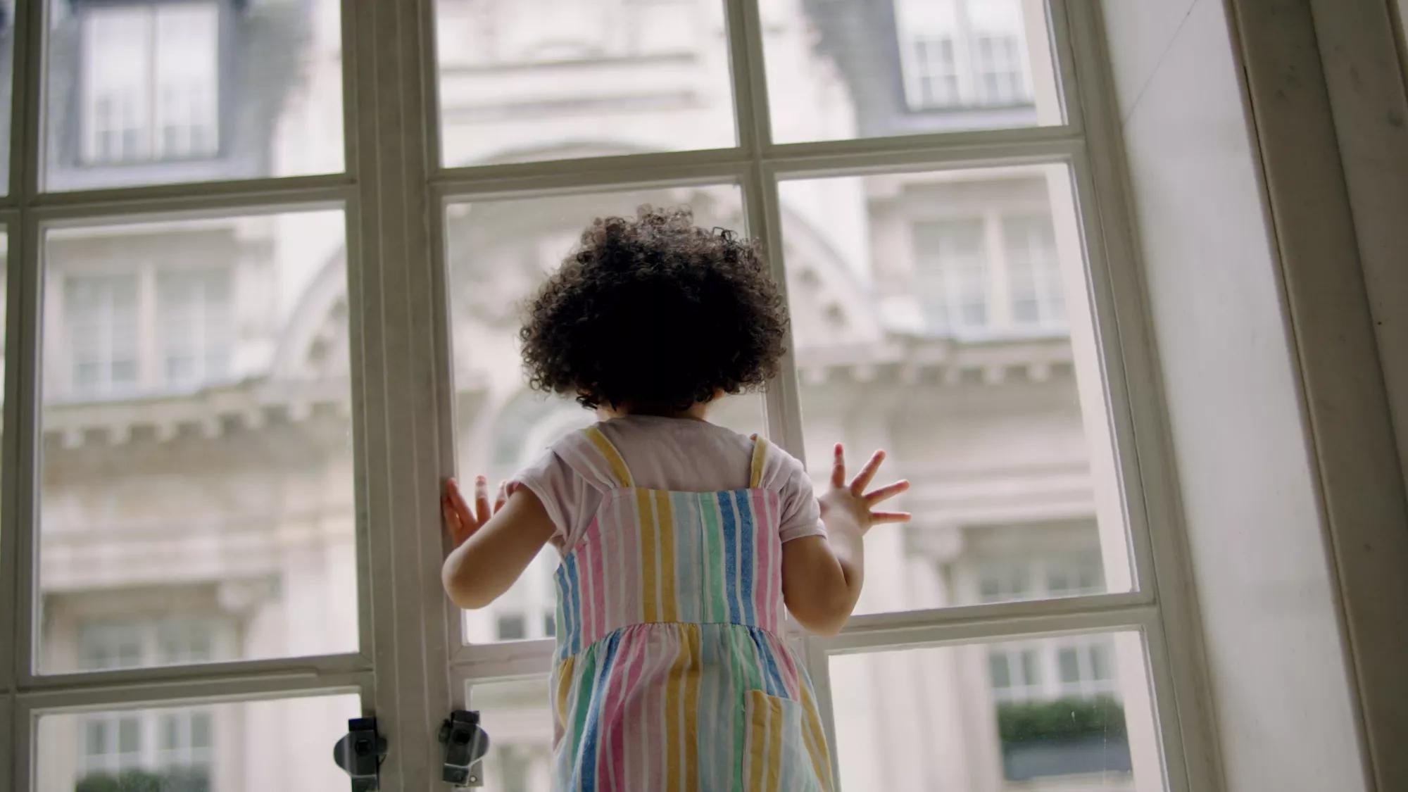 Child standing at a window looking out over London buildings in soft natural light