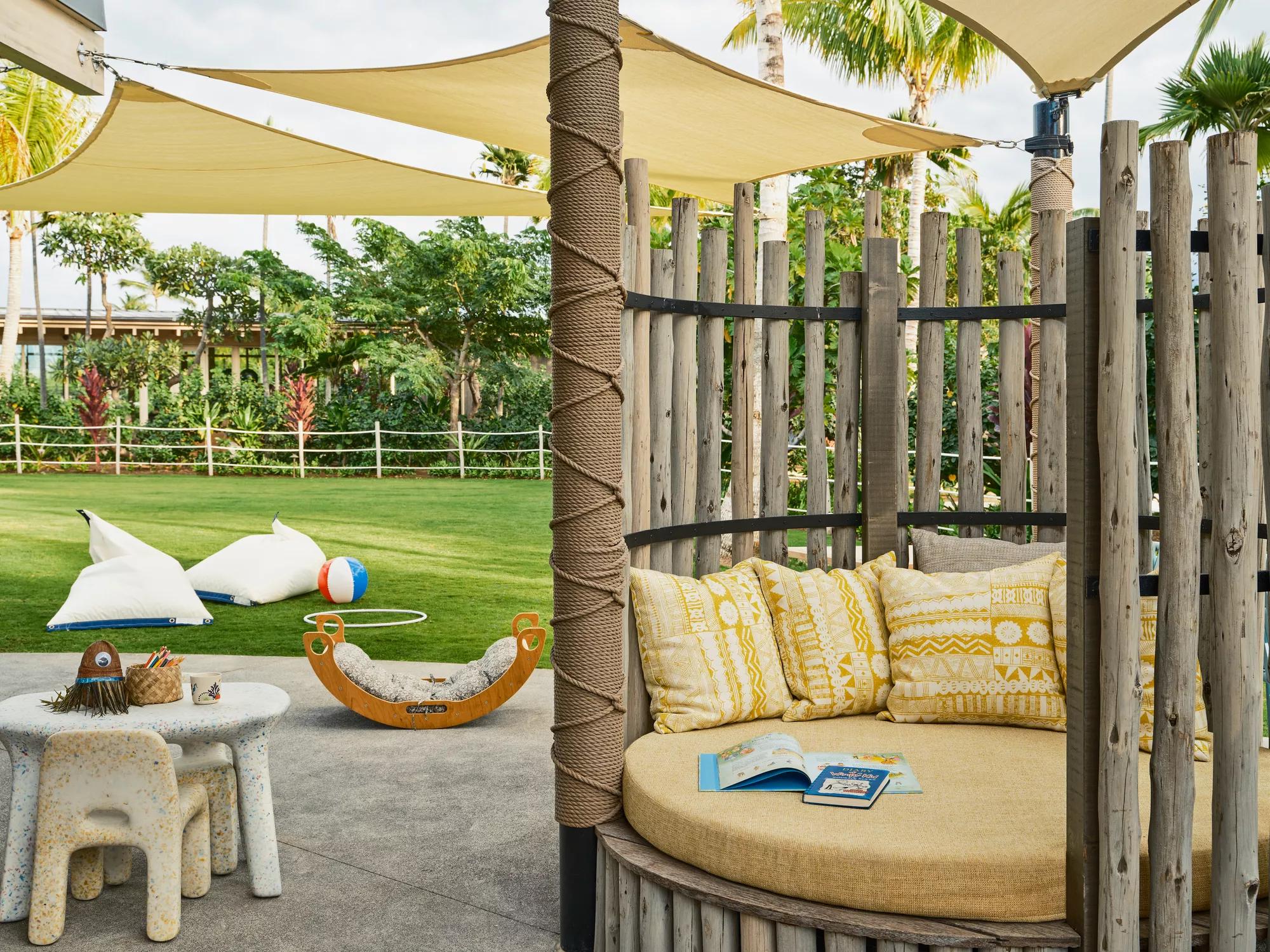 A grassy yard with children’s toys and a small table and chairs and a yellow daybed with cushions and books.