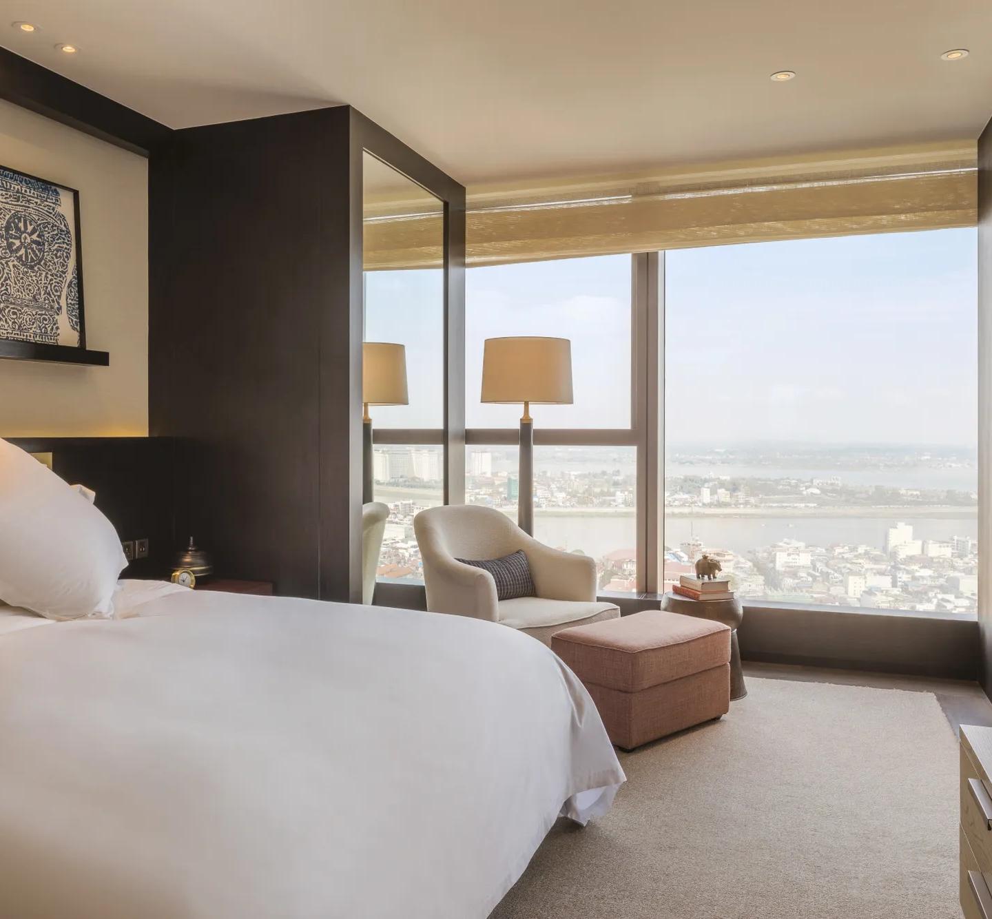 A modern bedroom with white linens, framed artwork above the bed, large windows overlooking a city and river, and a cozy seating area with an armchair, ottoman, and floor lamp.