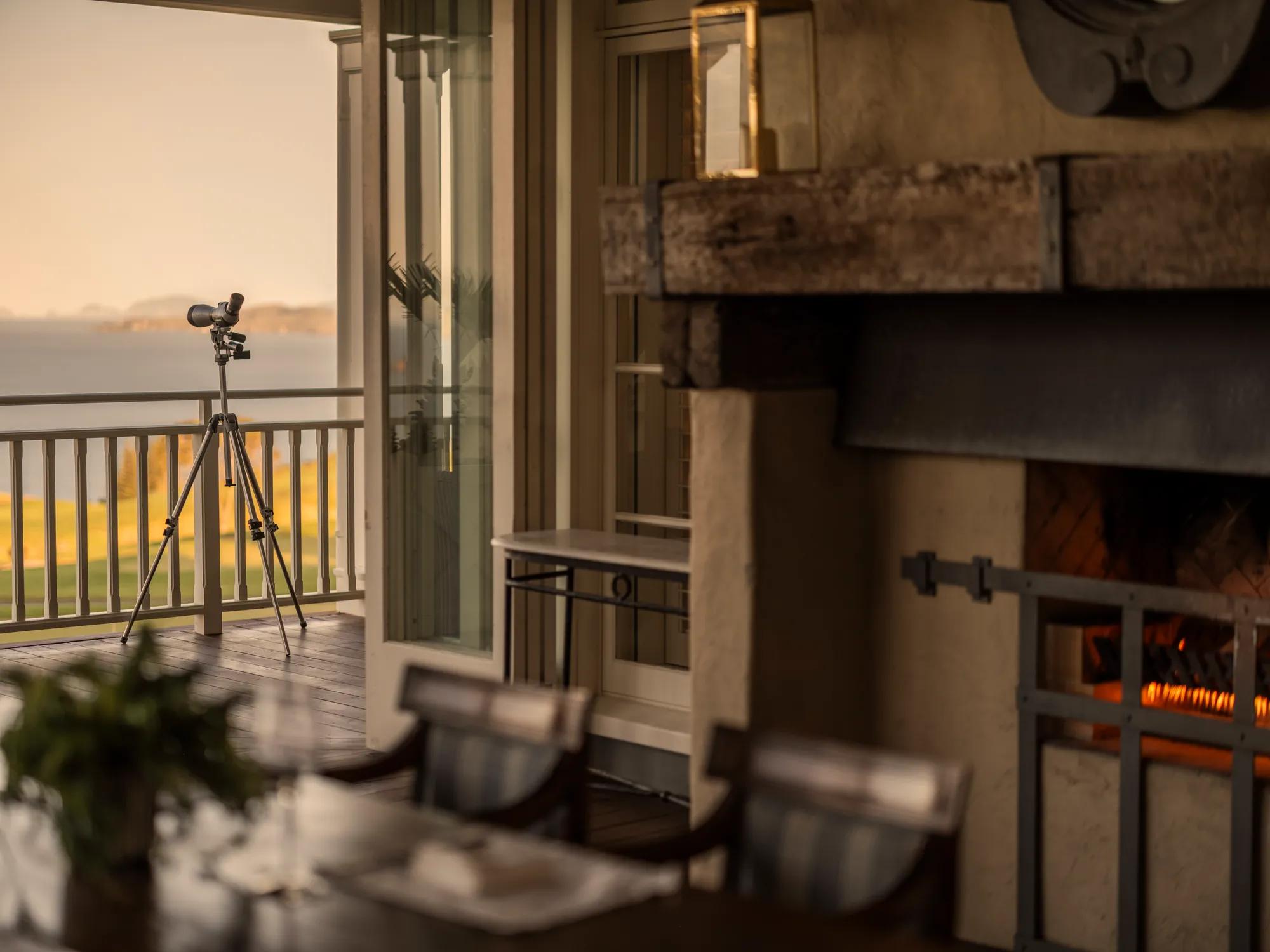 A covered outdoor terrace with wooden table and chairs in the foreground, a fireplace in the middle ground, and a balcony and telescope in the background, looking out to an ocean view.