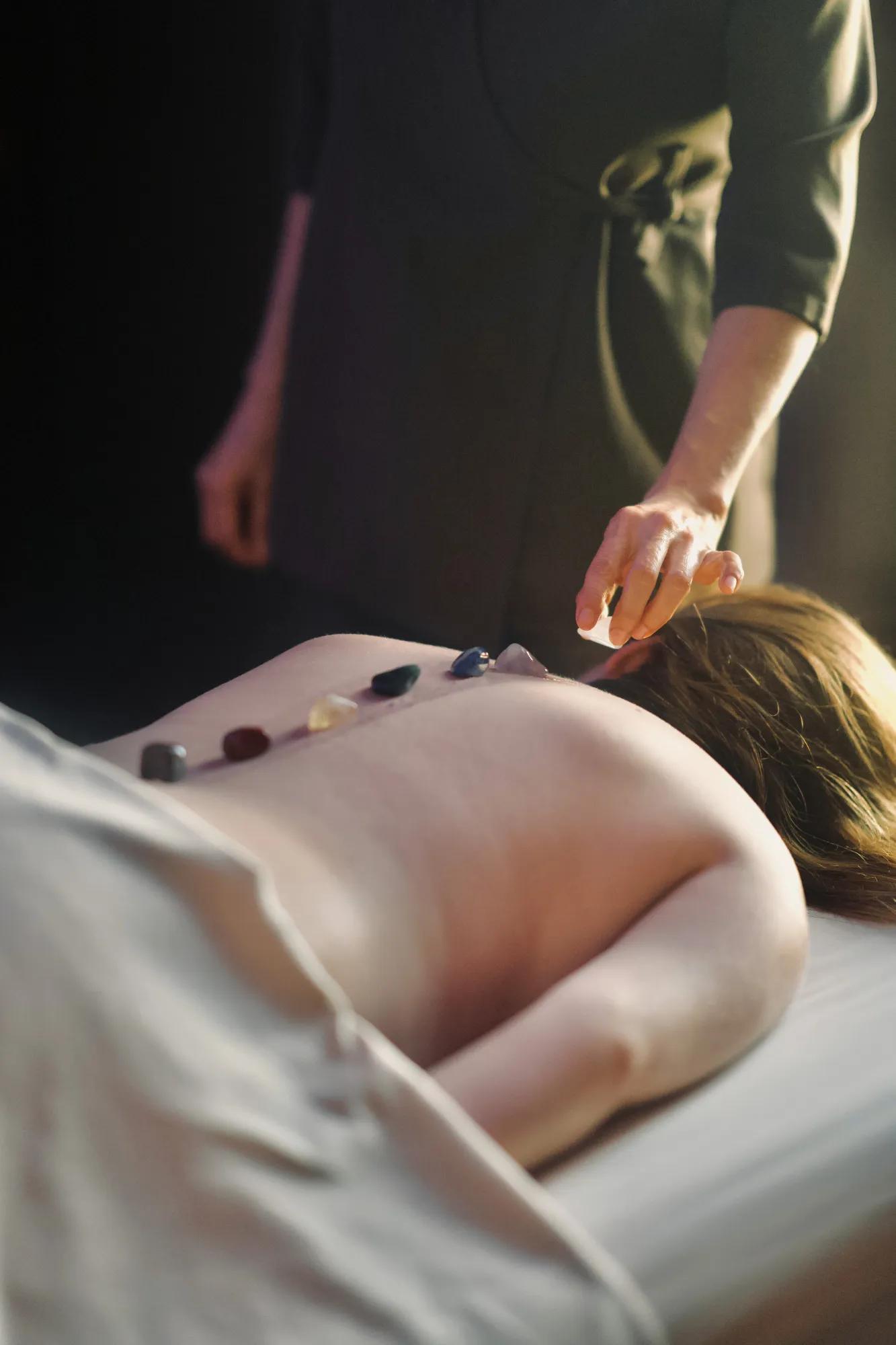 A woman lying face down on a massage bed with several colorful crystals placed along her spine, while a therapist adjusts them during a healing session.