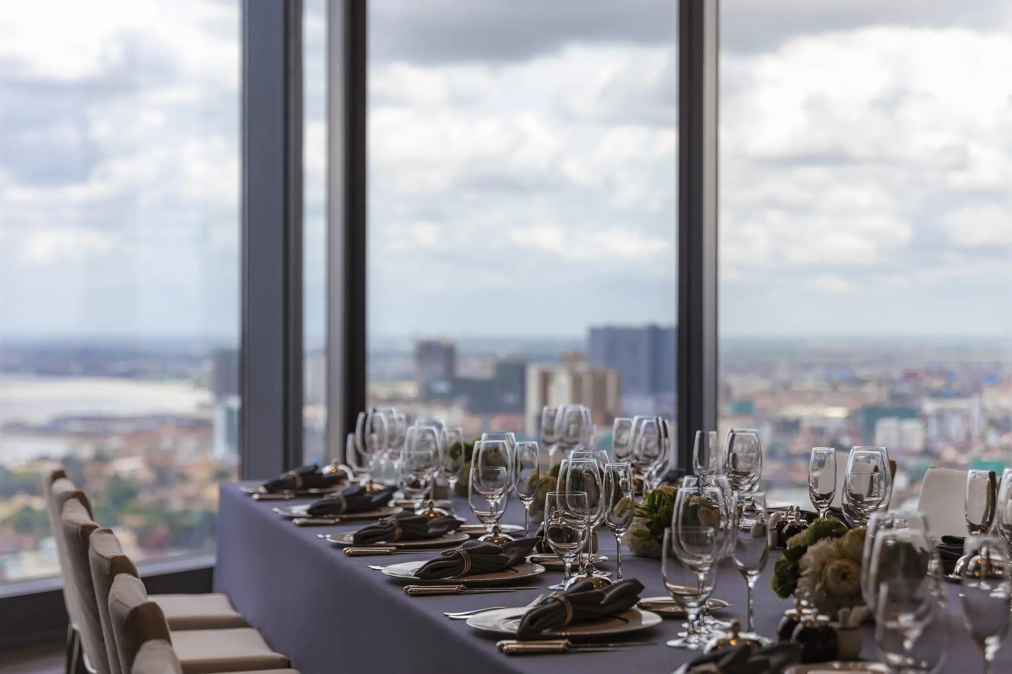 A formal dining table with a dark tablecloth, floral arrangements, and neatly arranged wine glasses, plates, and cutlery, set against large windows showcasing a panoramic city view