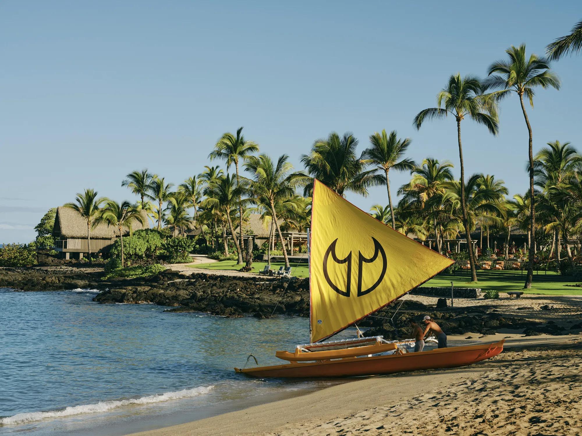 Two individuals pushing a Hawaiian sailing canoe into the ocean with a yellow sail and two black crab claw sail symbols on the front