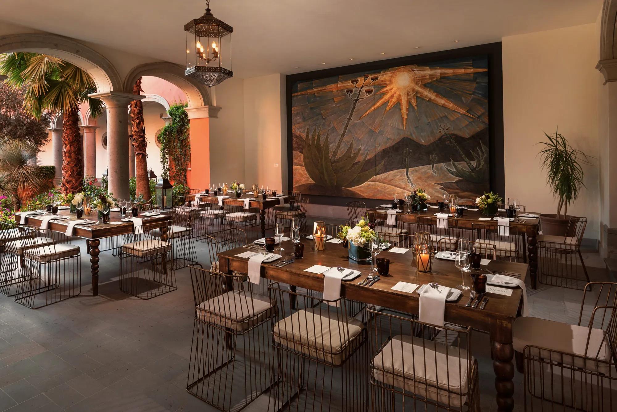 Semi-open event space with four round tables set for eight guests each. The covered area opens onto a garden on the left, featuring a chandelier overhead and a large framed artwork on the back wall.