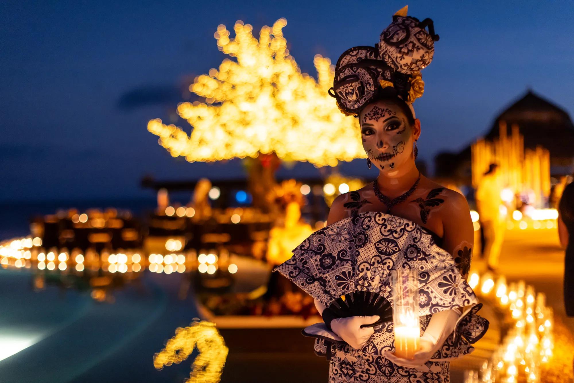 A woman in traditional folkloric dress dances gracefully, holding a decorative fan and lifting her lace skirt, under glowing golden lights. A man in a white outfit and hat stands smiling in the background, creating a festive Día de Muertos atmosphere.