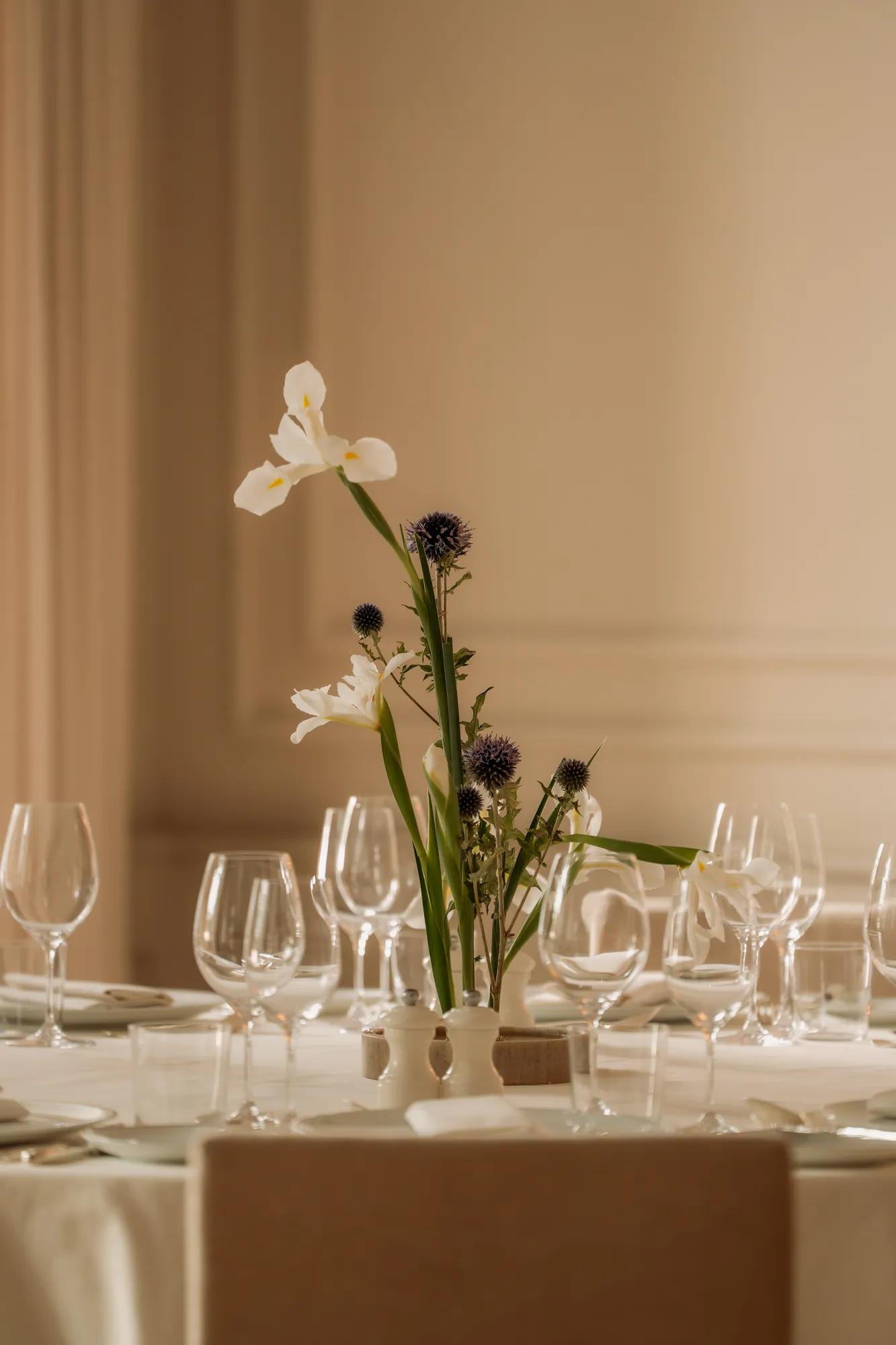 A tabletop with white linens, topped with glassware, plates and a vase of white flowers and thistle. 