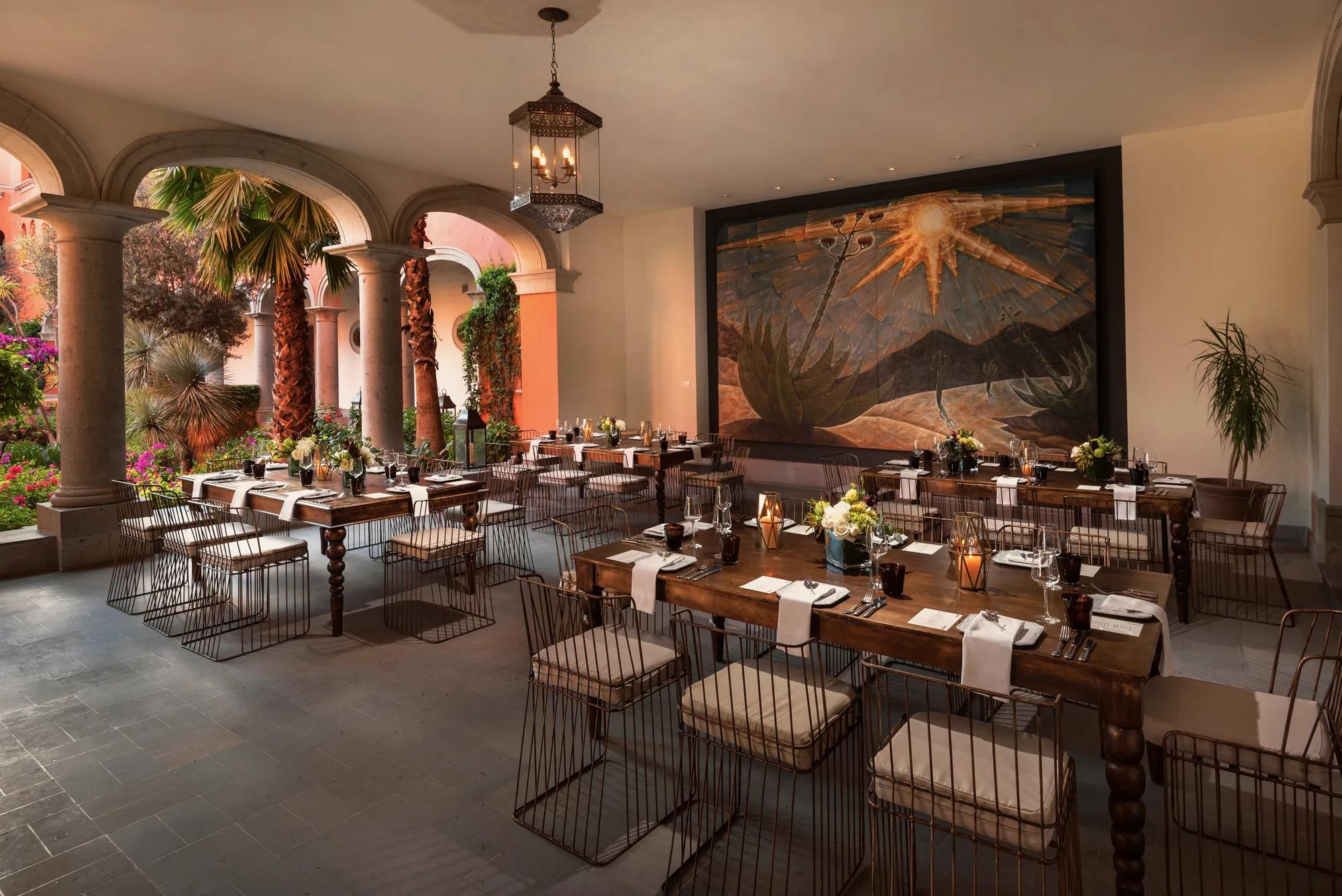 Elegant event space with several rectangular tables set for six to eight guests, featuring a large artwork on the back wall and a semi-open layout with garden views.